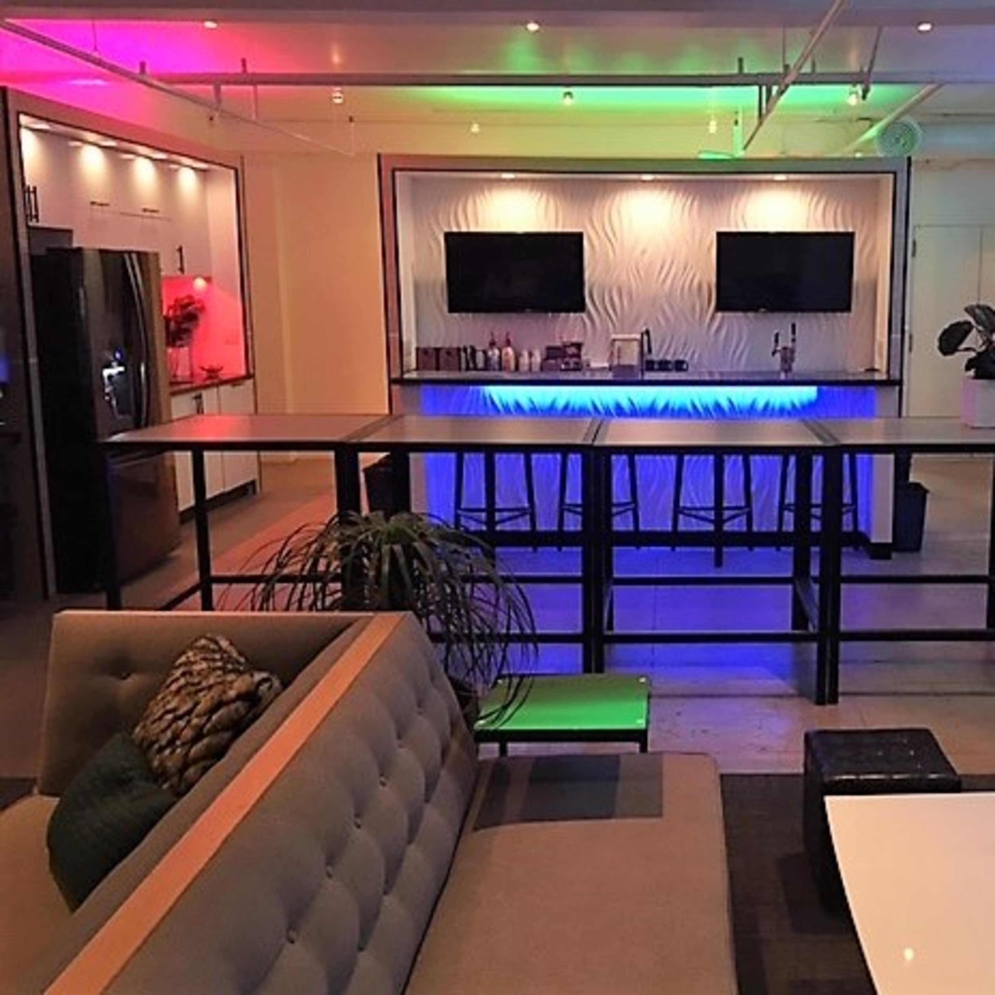 The image shows a modern lounge area with a bar featuring illuminated counters, two television screens, and a seating arrangement nearby.