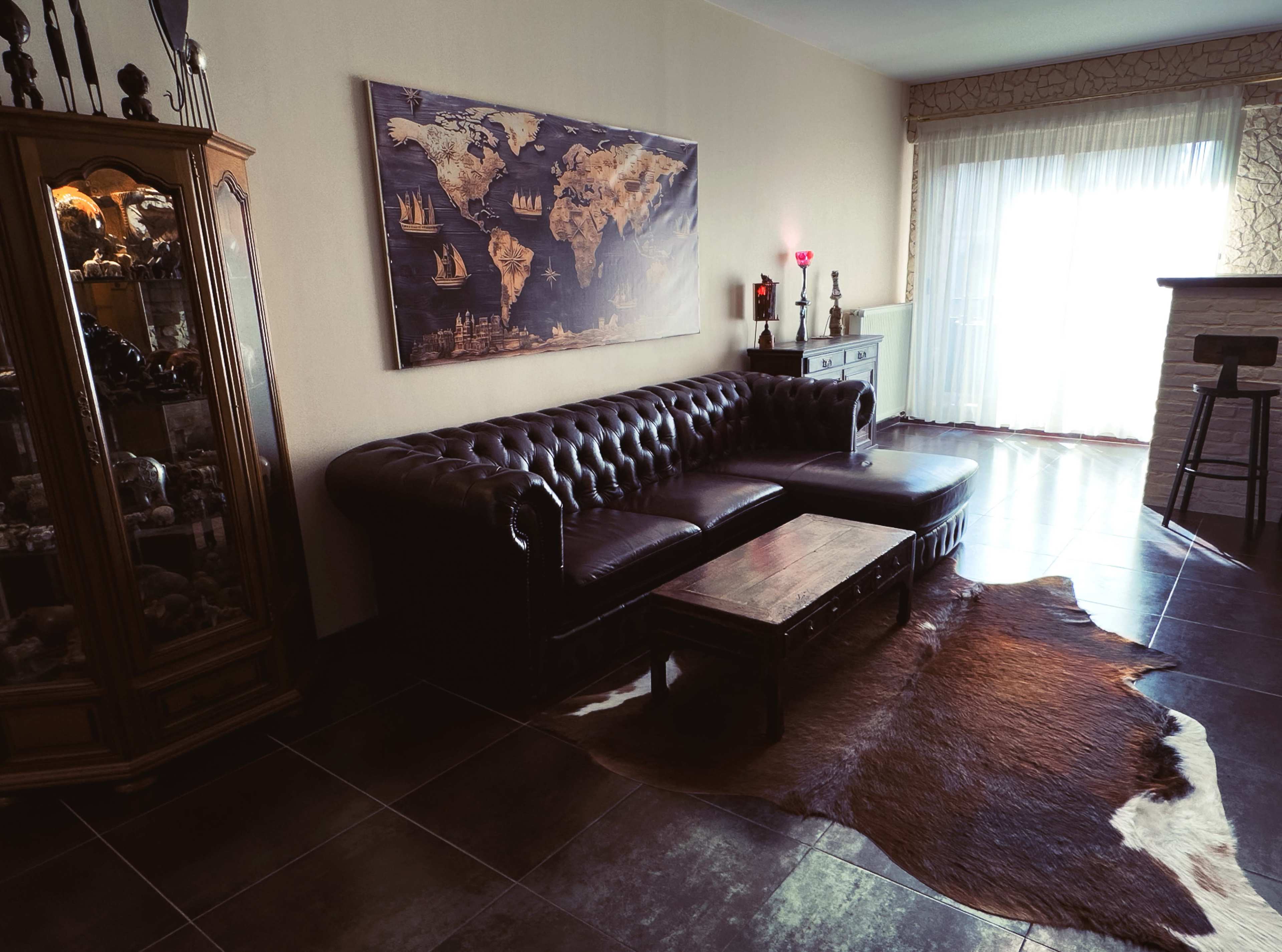 The image shows a living room with a dark leather sectional sofa, a wooden coffee table, a world map artwork on the wall, and a large window with sheer curtains.