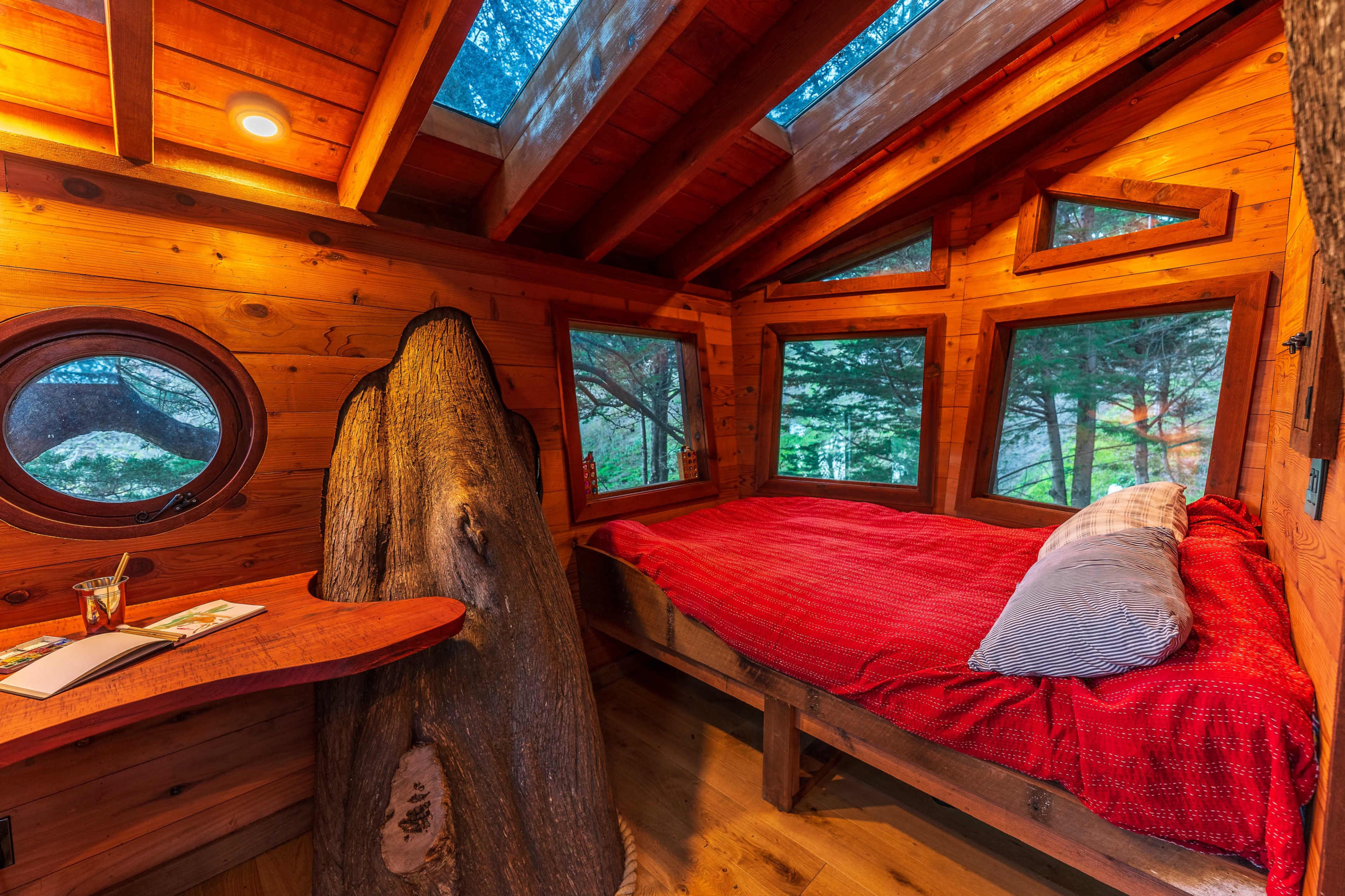 The cozy interior of a wooden cabin features a bed with a red blanket, large windows, and a small table made from a log.