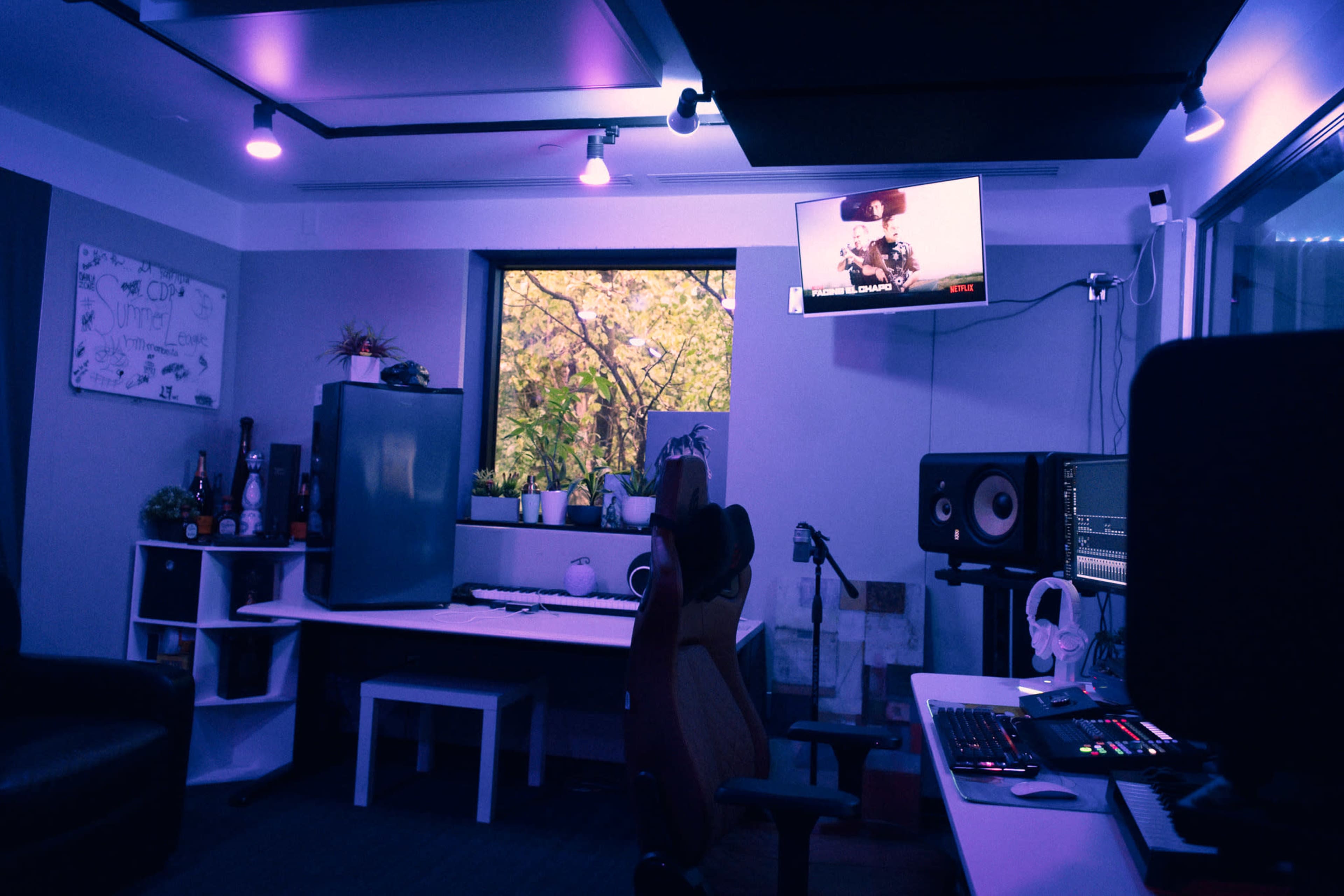 A modern music studio features a desk with electronic equipment, a television displaying content, and a large window showing outdoor greenery, all illuminated by purple lighting.