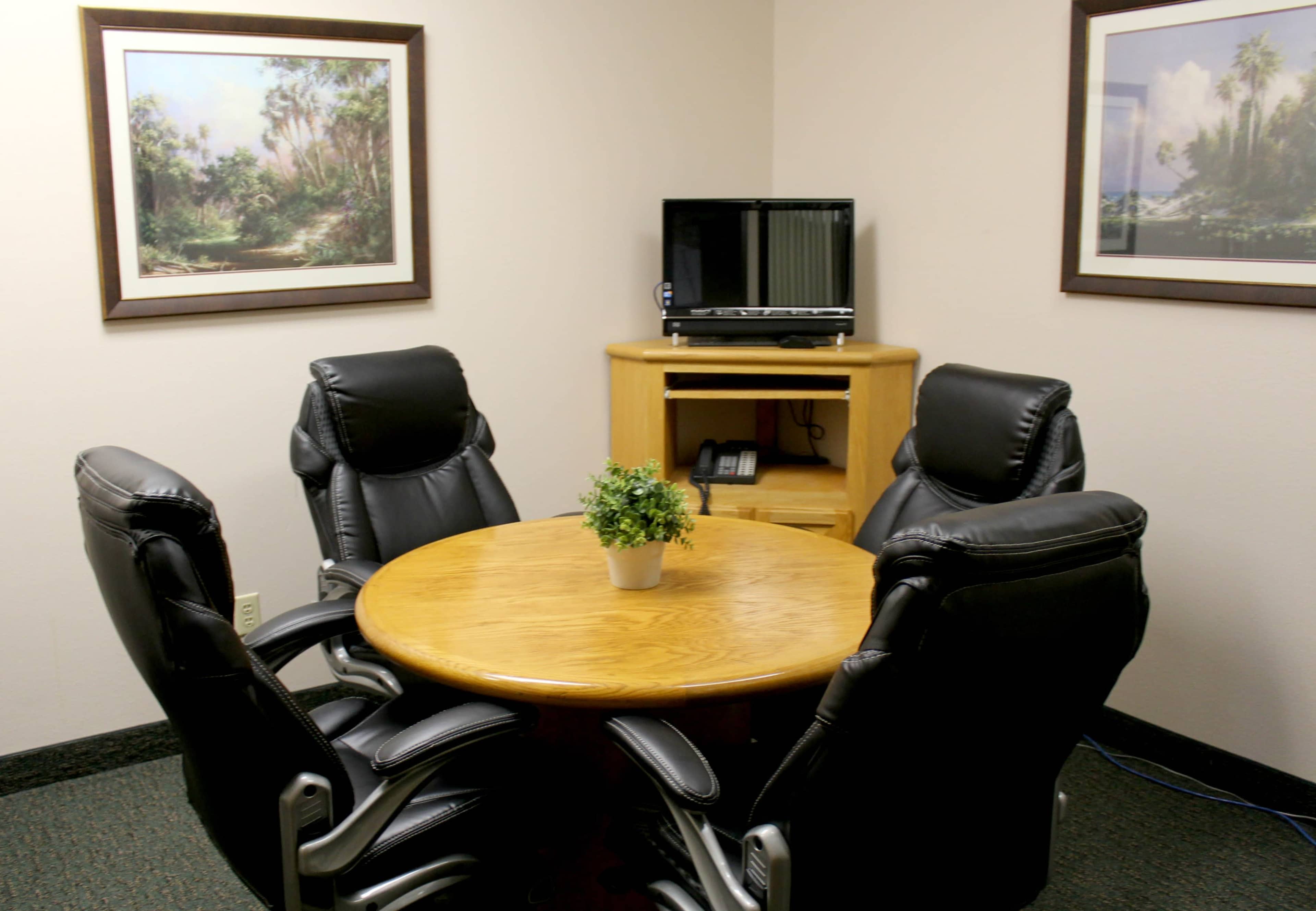 A small conference room features a wooden round table surrounded by four black leather chairs, with a television and a potted plant on a nearby cabinet.