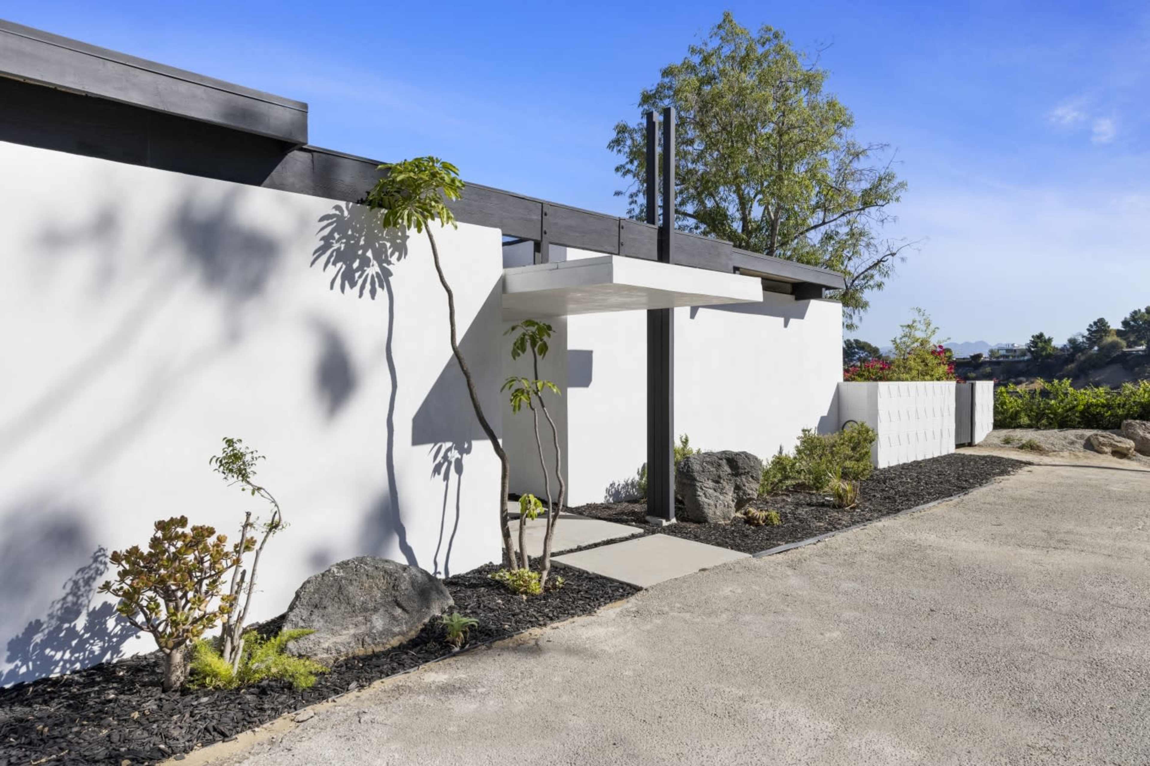The image shows a modern white house with a flat roof, featuring a covered entrance, surrounded by landscaped greenery and rocky elements.
