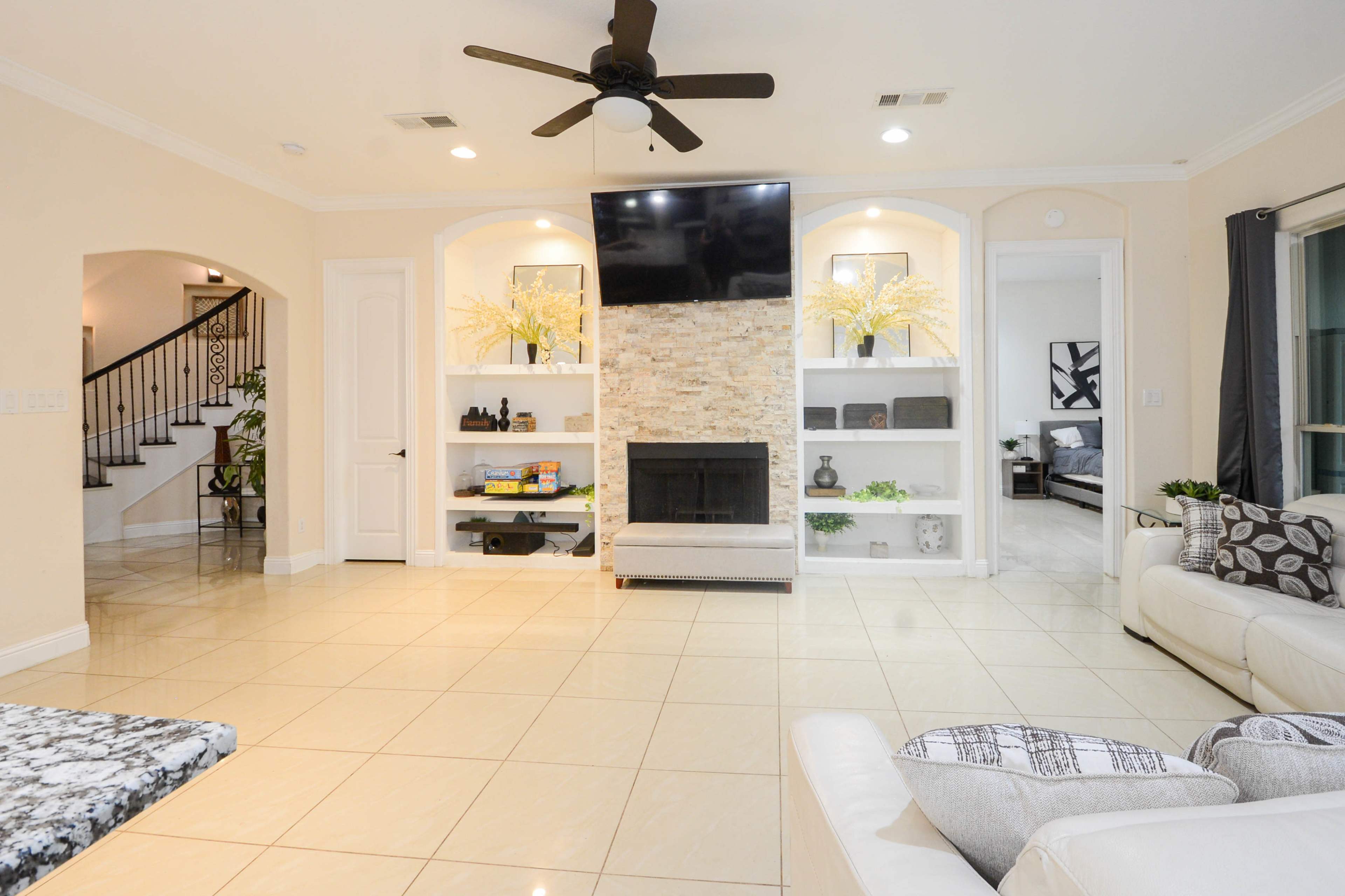 A spacious living room features a stone fireplace with built-in shelves and a large wall-mounted TV, surrounded by light-colored walls and tiled flooring.