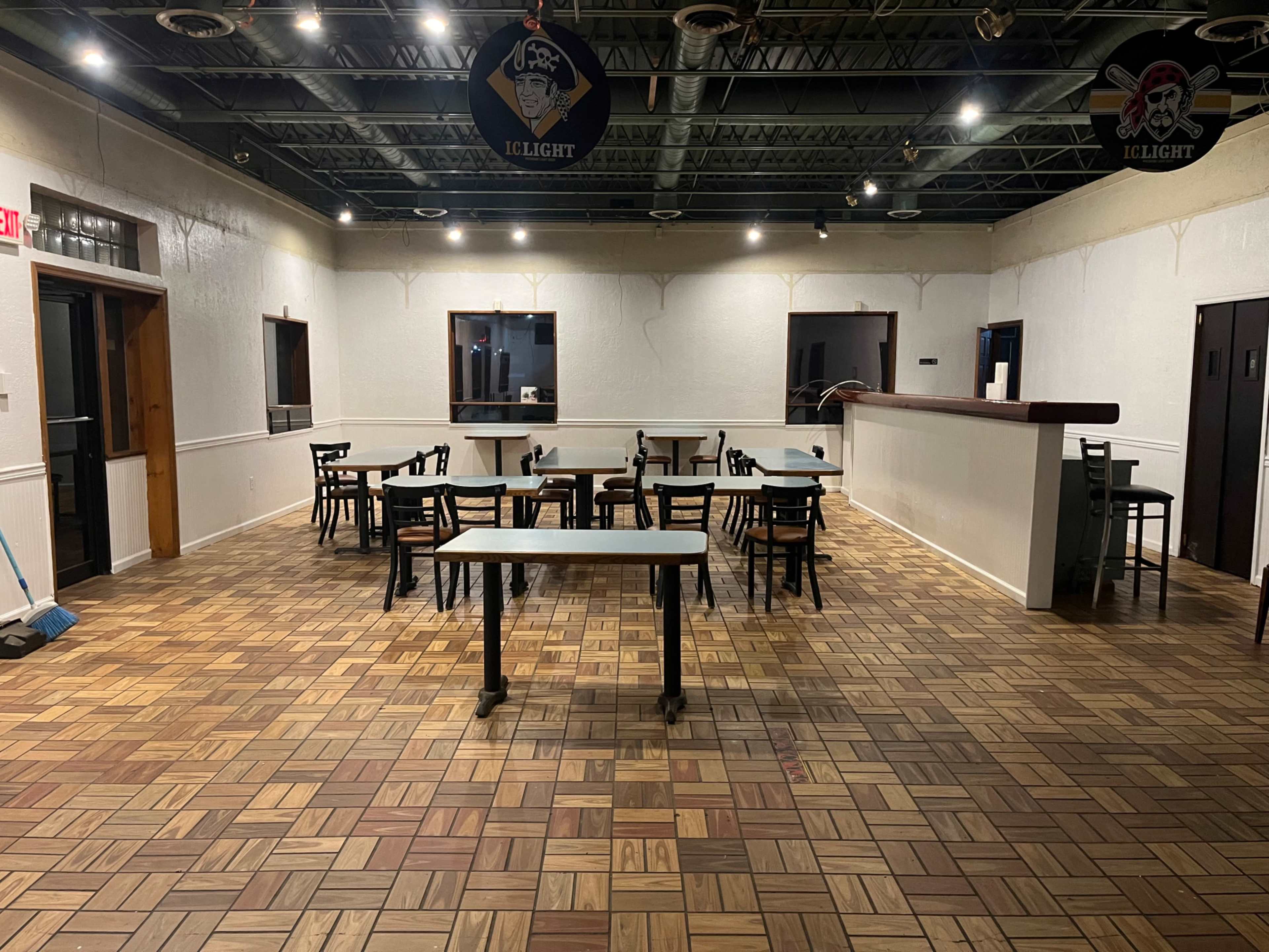 An empty room features several tables and chairs arranged on a patterned wooden floor, with walls painted white and a bar area in the back.