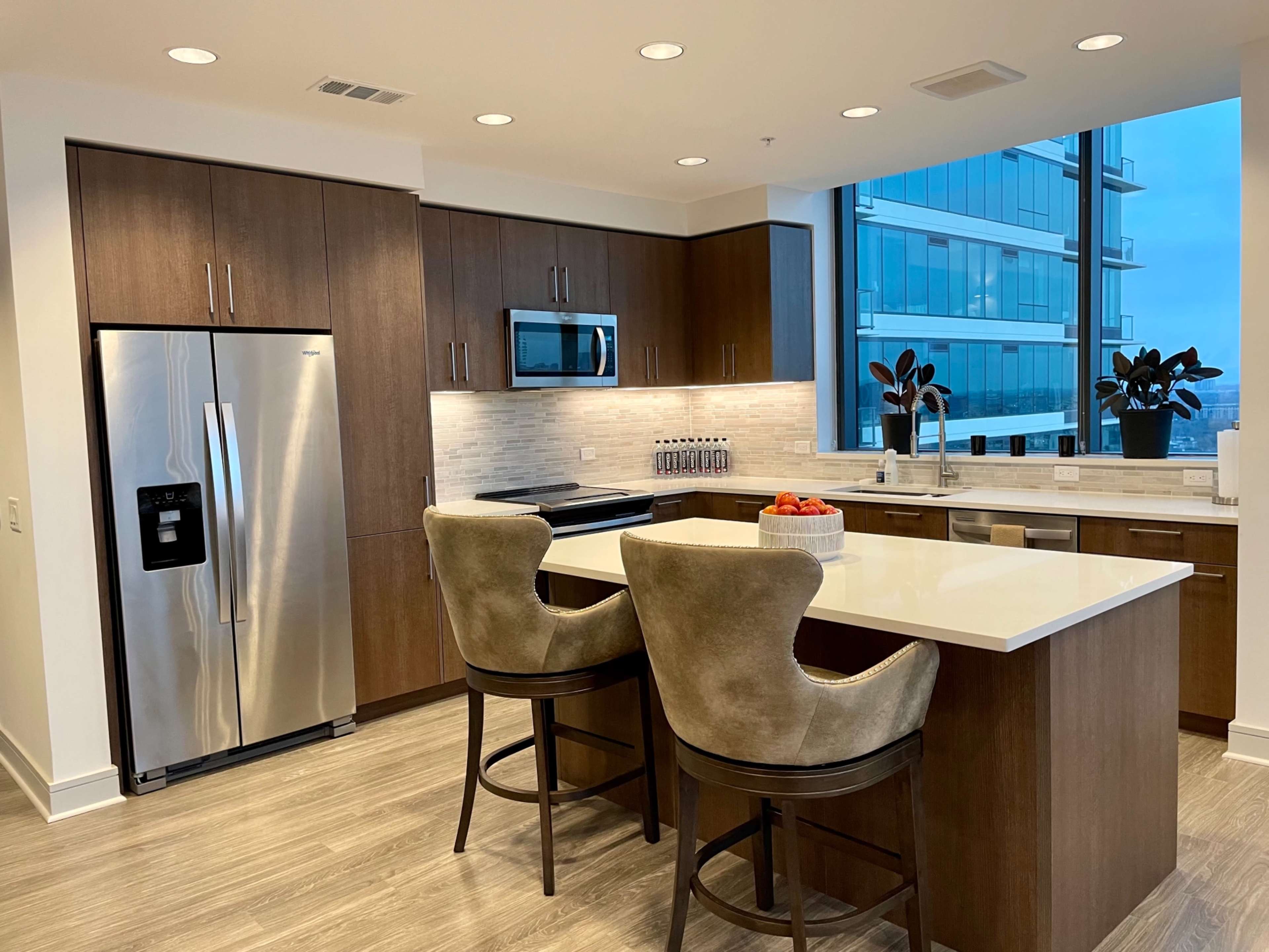 The kitchen features dark wood cabinetry, stainless steel appliances, a large white countertop, and two upholstered bar stools at the island.