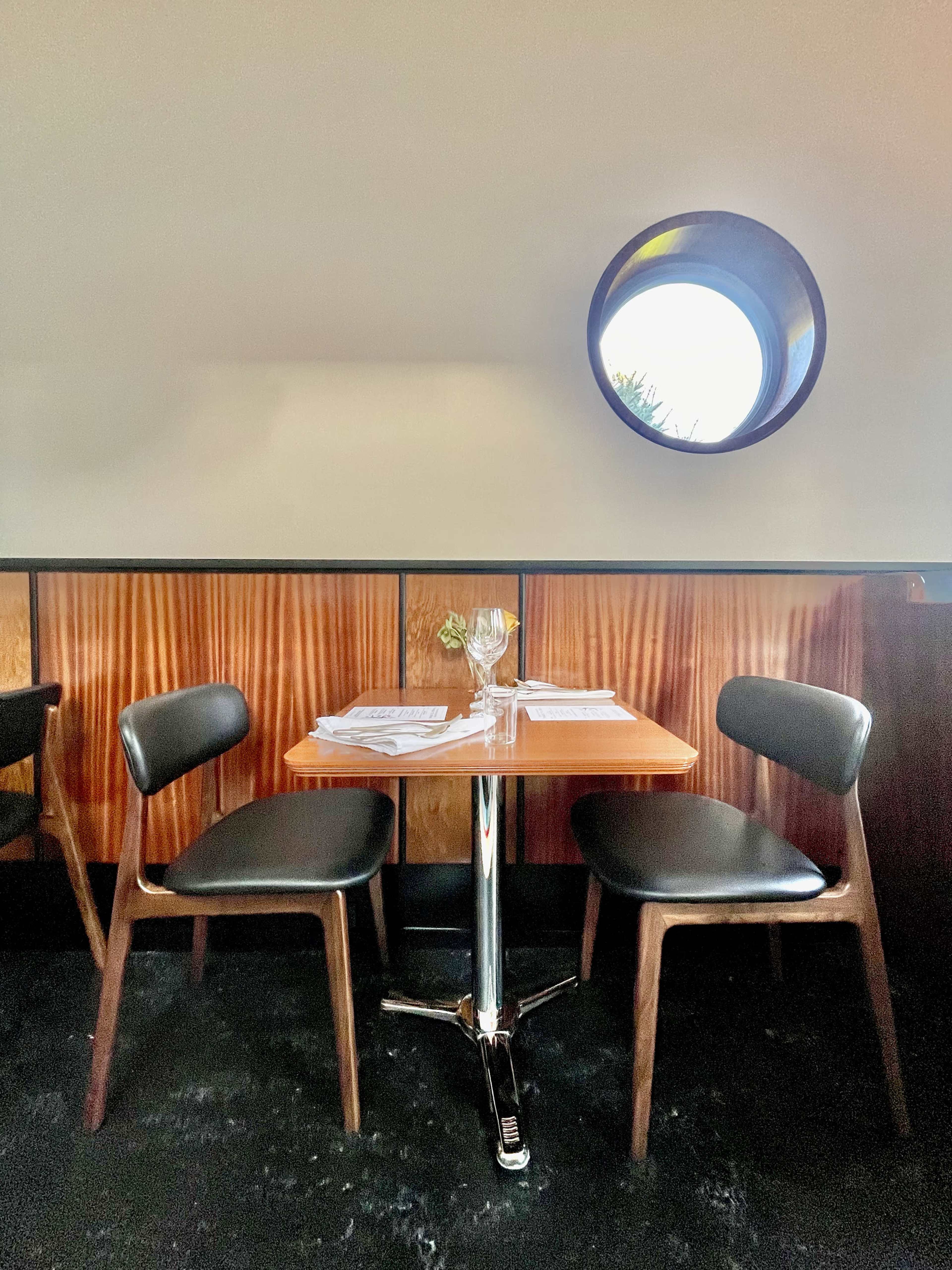 A small table with two chairs is set up against a wooden wall, featuring a round window above.