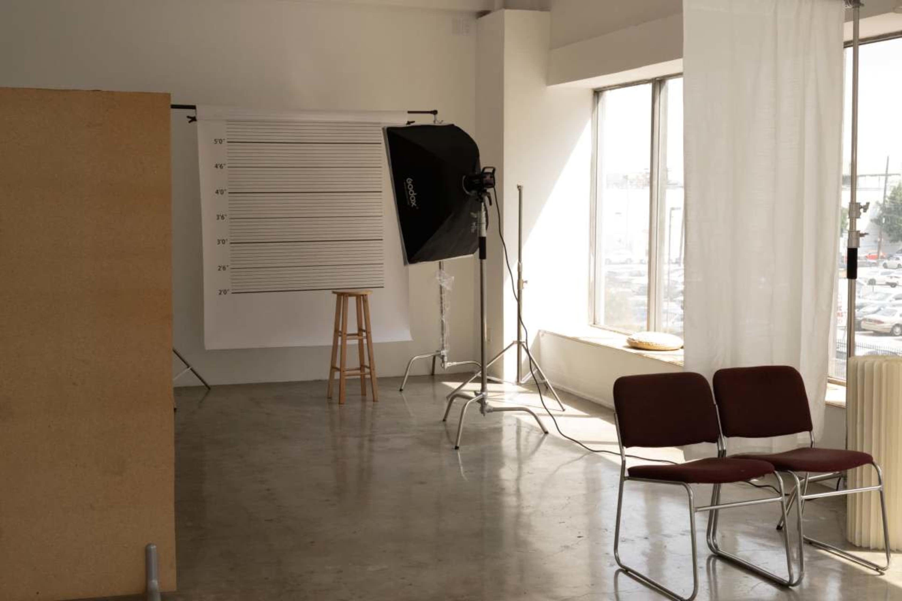 The image shows a spacious photo studio with a backdrop, lighting equipment, a stool, and two chairs arranged near a large window.