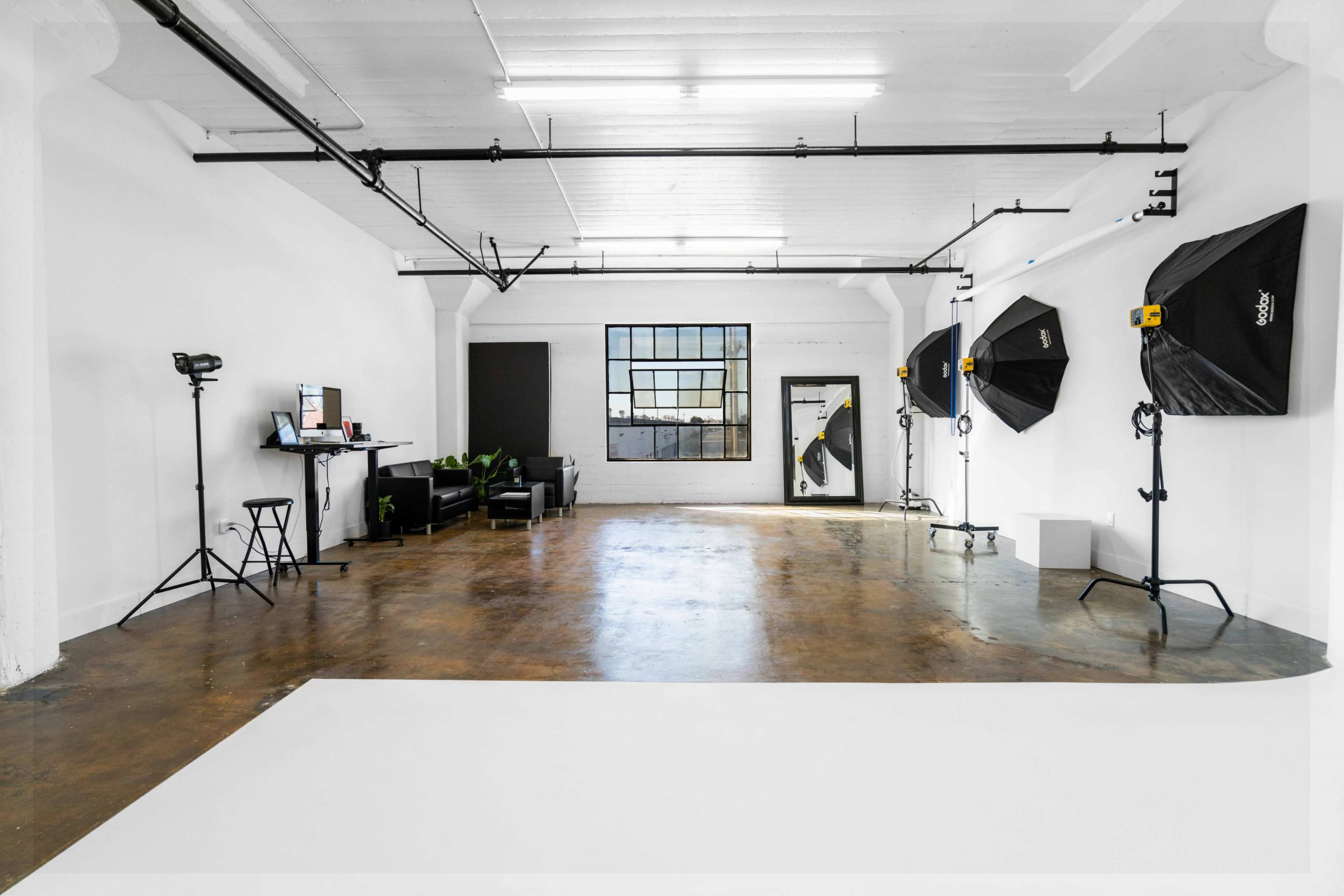 A spacious studio with a polished concrete floor, illuminated by softbox lights, a large window, and minimal furniture.