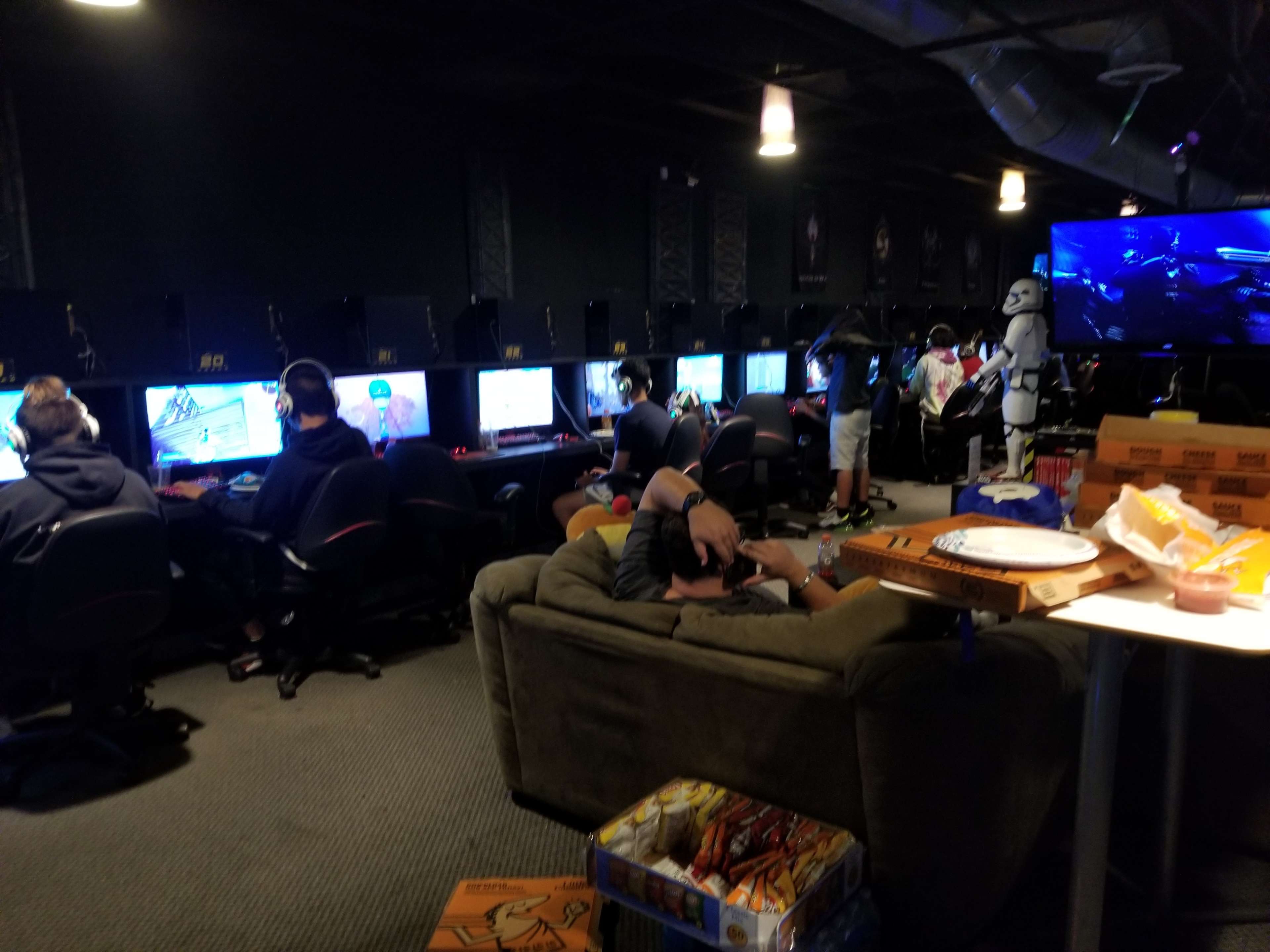 A gaming lounge with several players at computer stations, while others relax on a couch surrounded by pizza boxes and snacks.