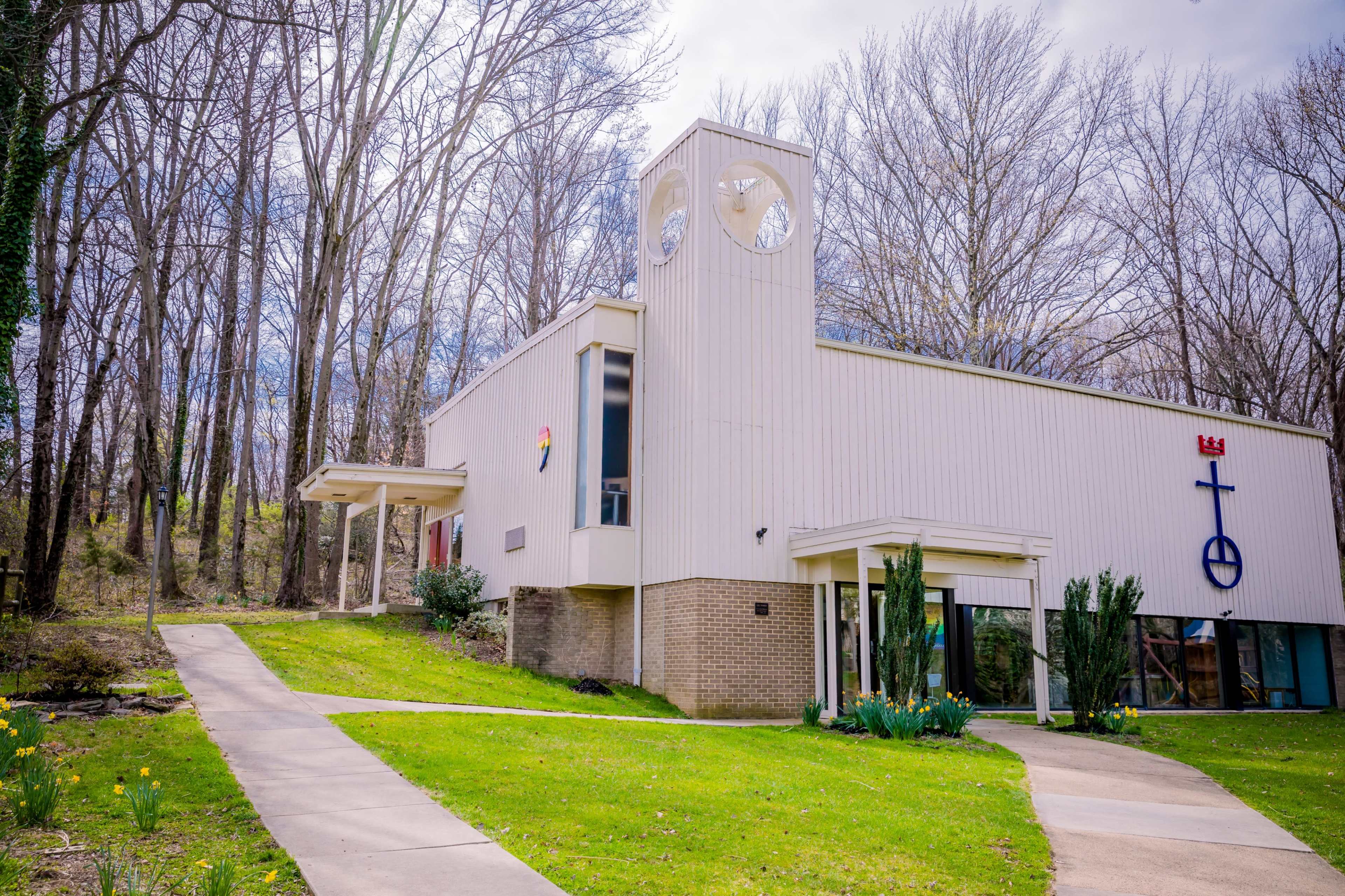 A modern building with a clock tower and religious symbols is surrounded by trees and a landscaped pathway.