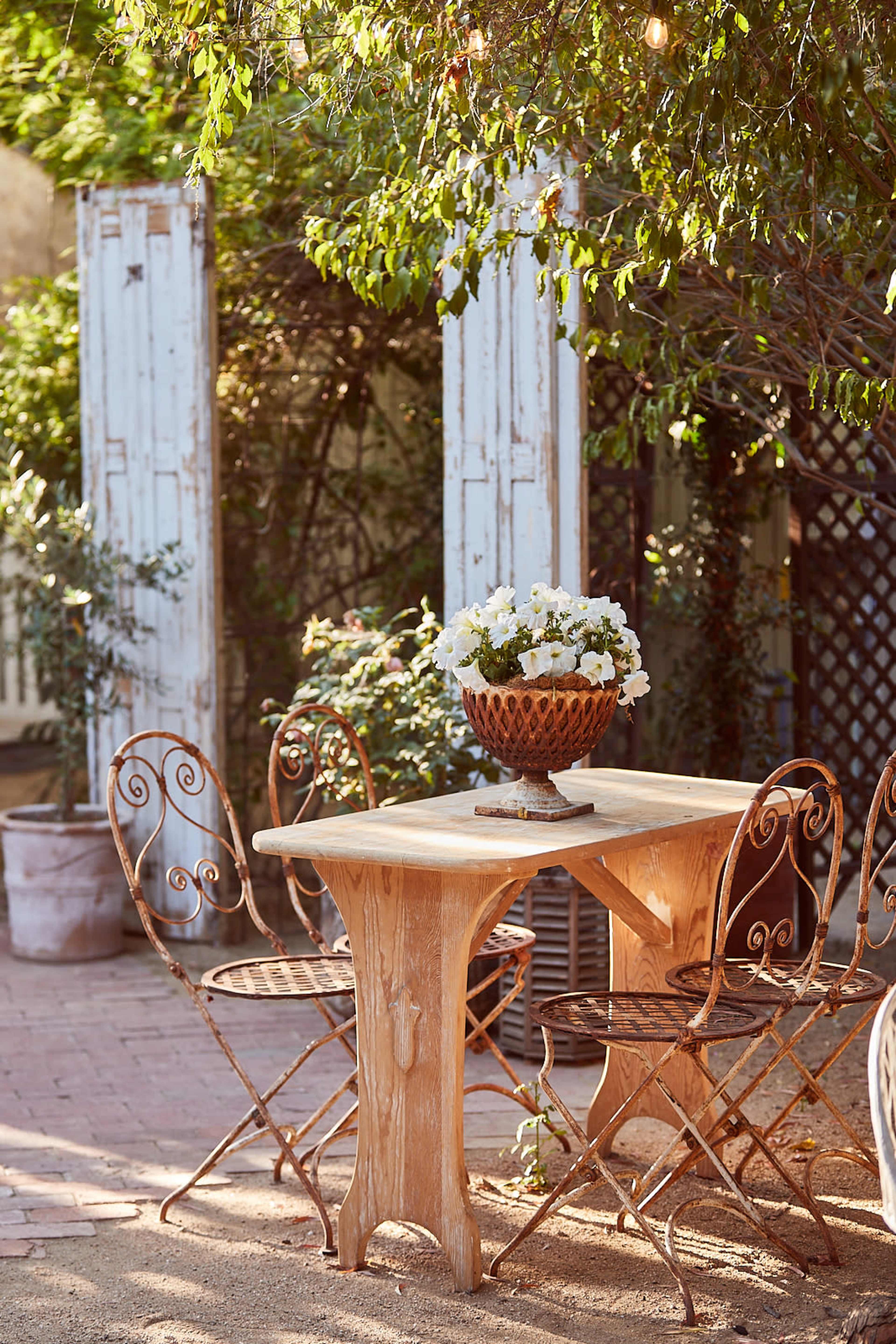 A rustic wooden table is set with a floral centerpiece, surrounded by ornate metal chairs in a garden setting.