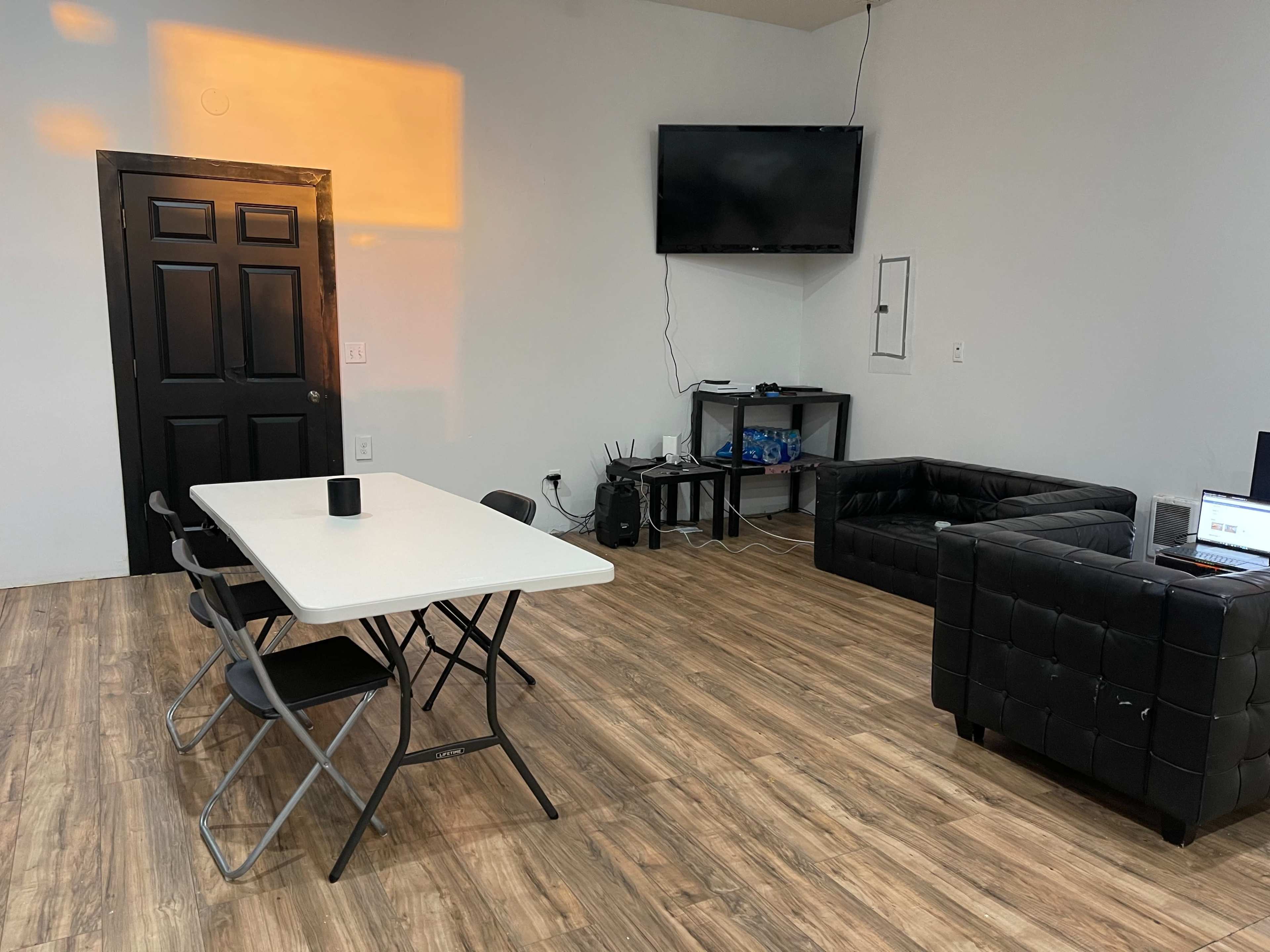 A simple room with a black table and chairs, a black leather couch, a television on the wall, and a small entertainment unit against the wall.