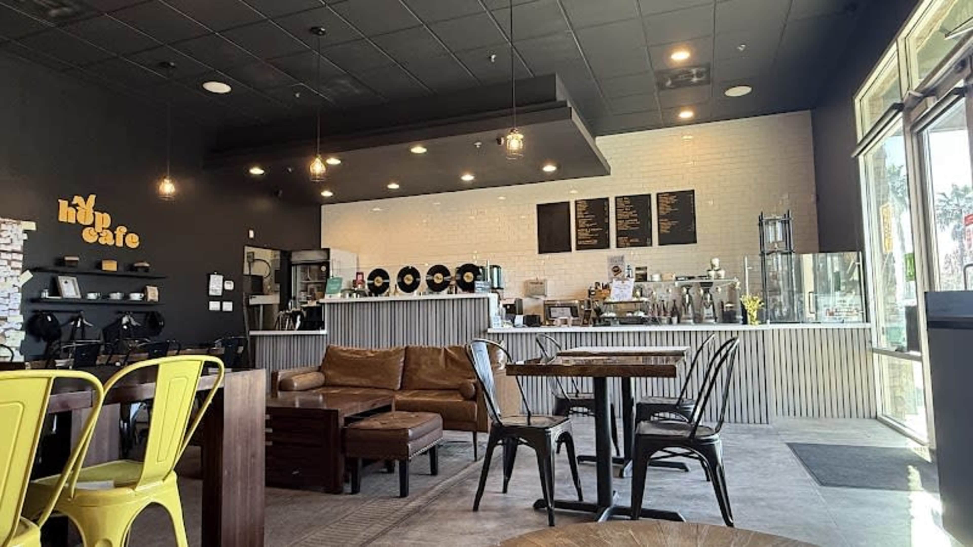 The image shows the interior of a café featuring a mix of seating options, a counter with coffee equipment, and decorative vinyl records on the wall.