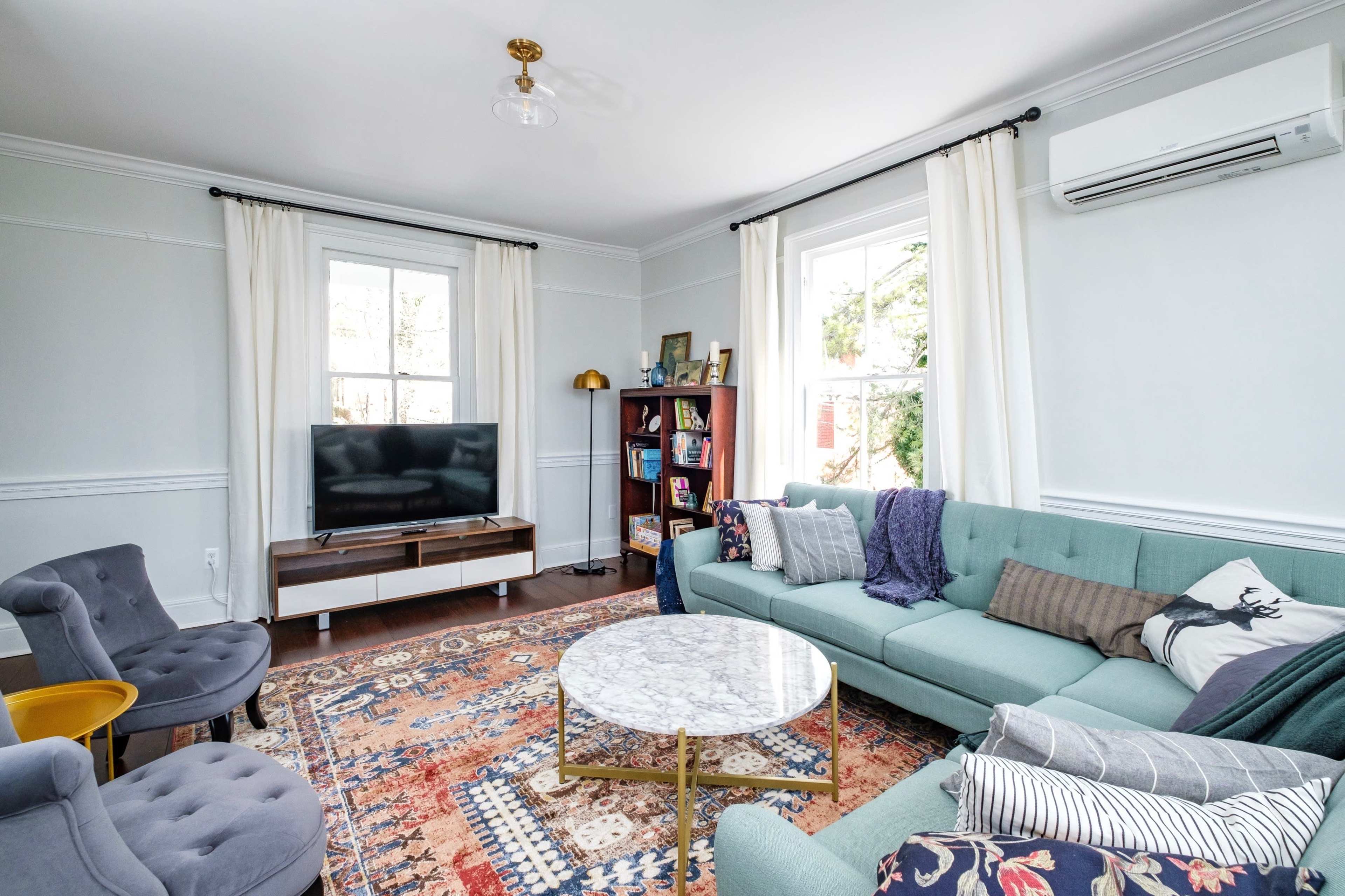 The room features a teal sectional sofa and armchairs, a round marble coffee table, a television on a console, and bookshelves filled with various items.