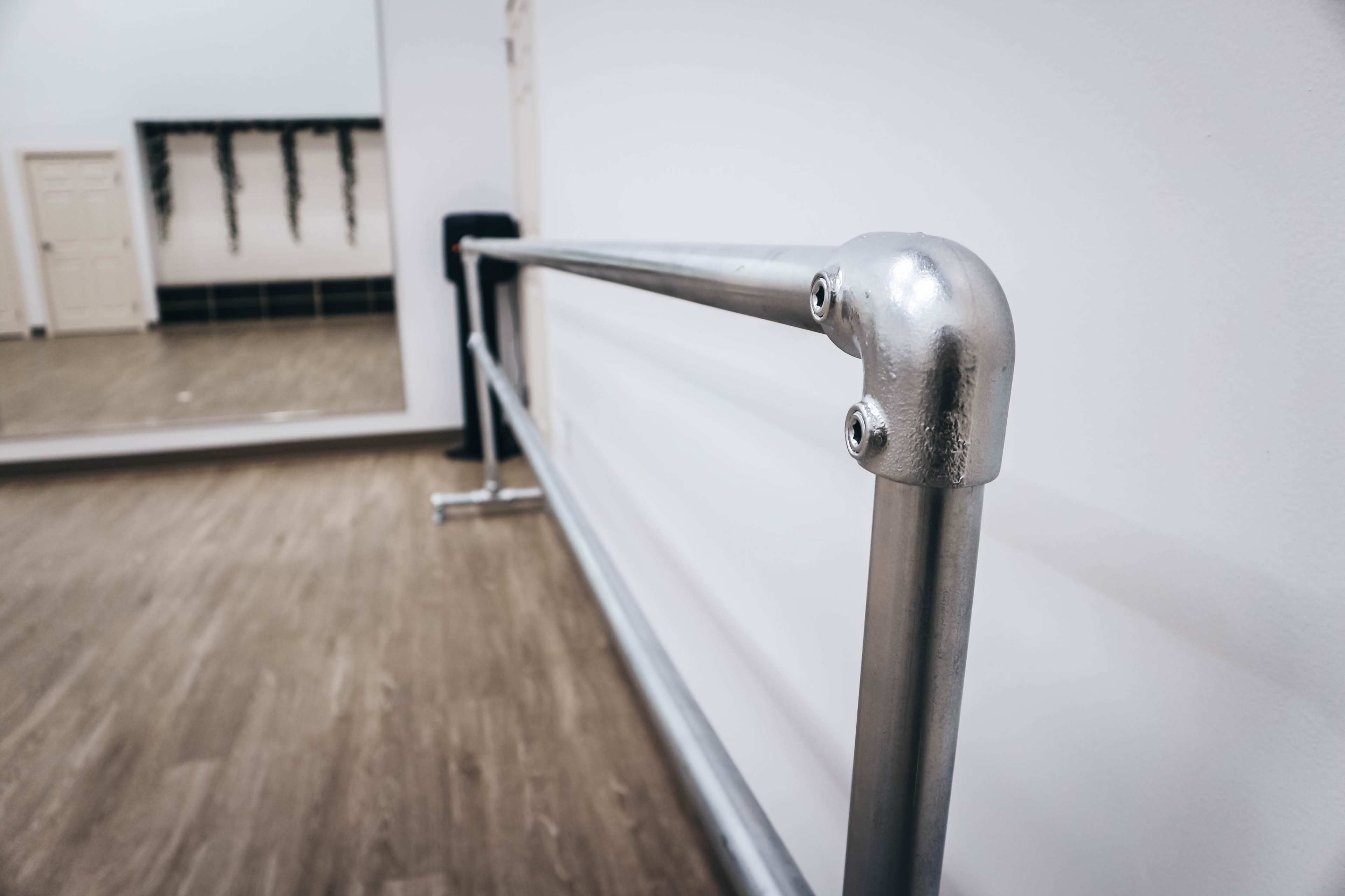 A metal ballet barre is mounted on a white wall in a mirrored dance studio.