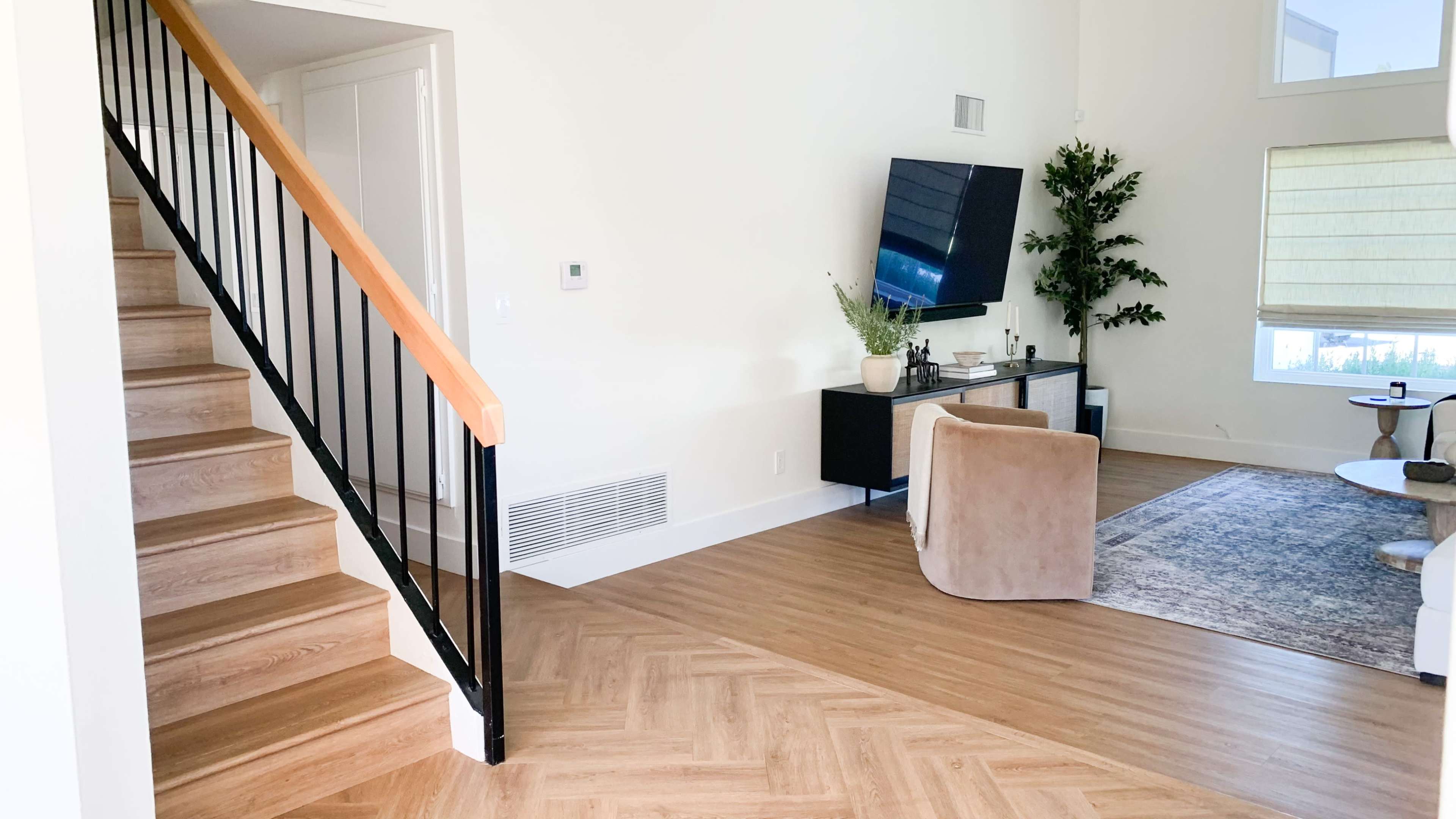 A modern living room with a staircase on the left, a television mounted on the wall, and a cozy seating area with a rug and decorative plants.
