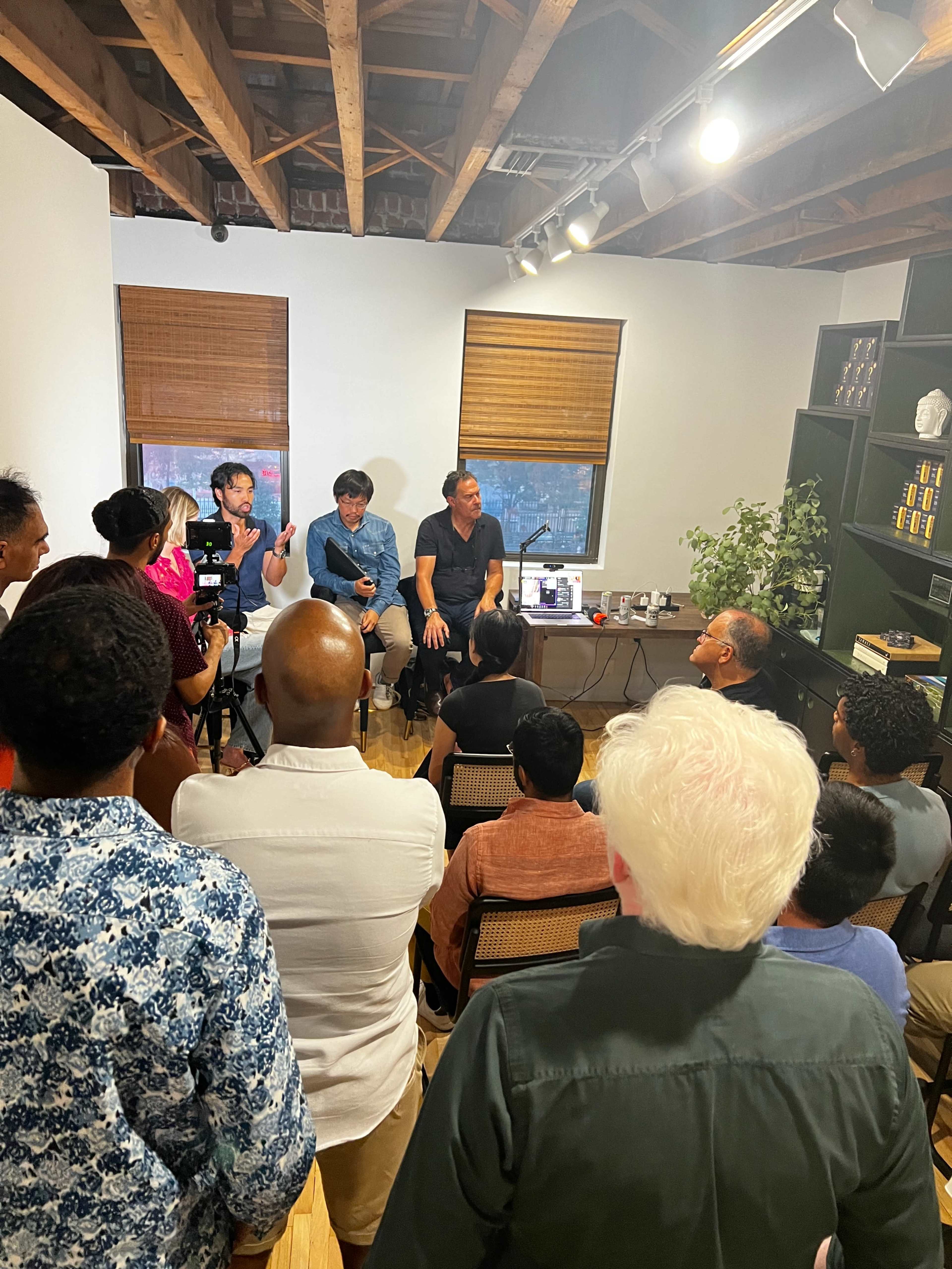A diverse group of people is seated, listening to two speakers engaged in a discussion in a well-lit indoor setting.