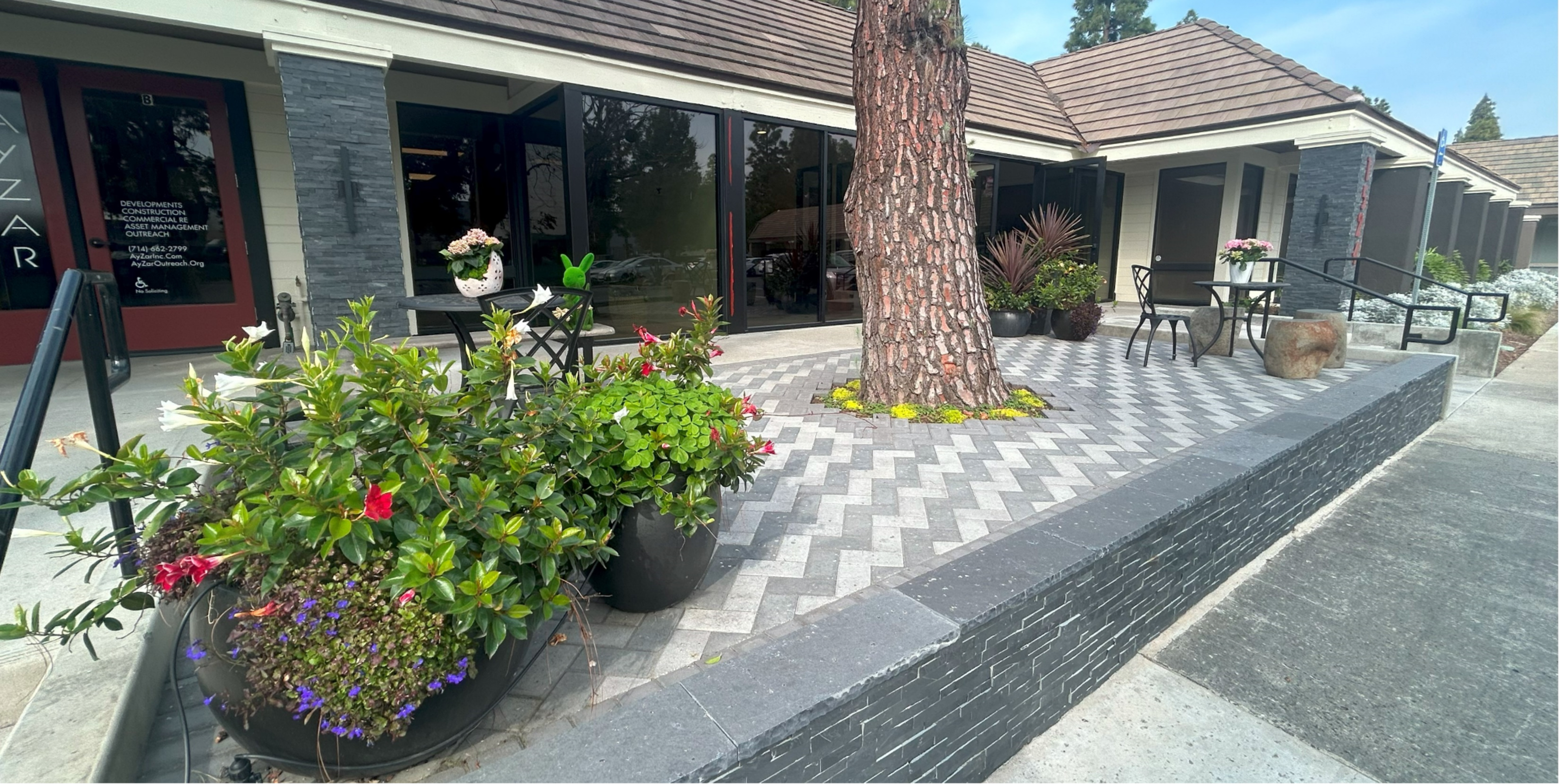 The image shows a landscaped area in front of a commercial building, featuring a patterned stone patio, potted plants, and a tree in the center.