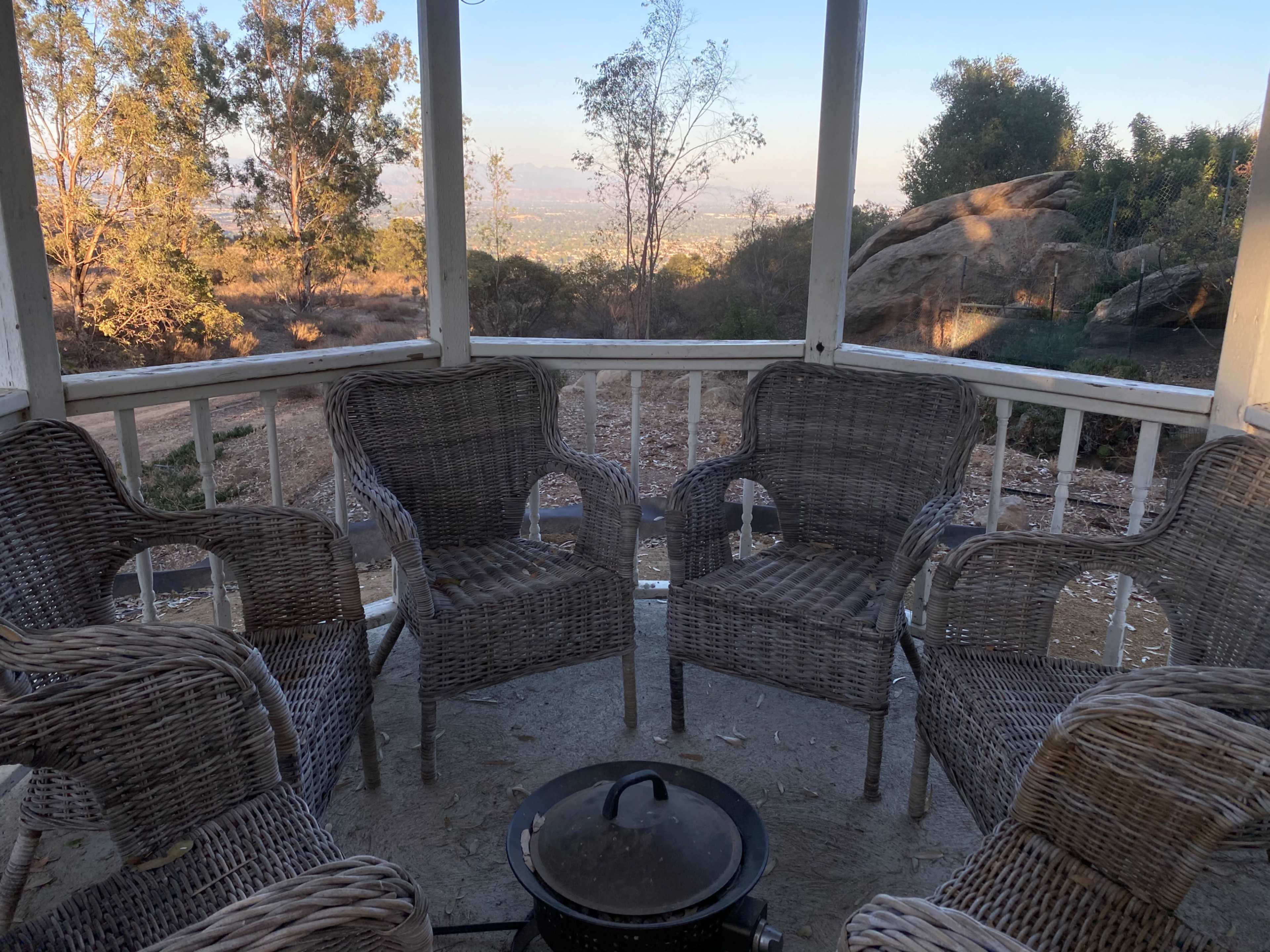 A circular patio with four wicker chairs surrounds a black pot in an open gazebo, overlooking a valley filled with trees and distant hills.
