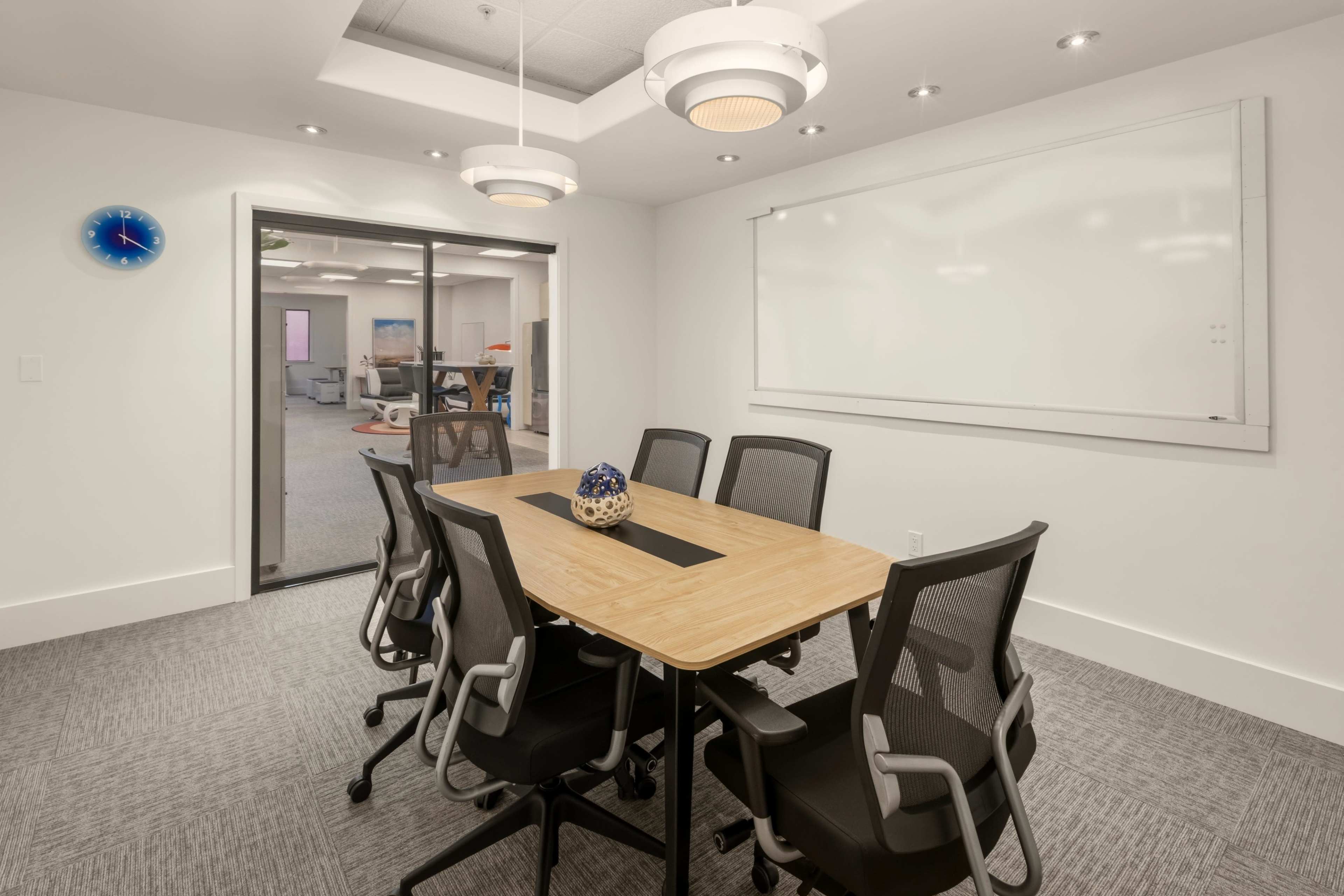 The image shows a modern conference room with a wooden table surrounded by six black chairs, a whiteboard on the wall, and a glass door leading to another room.