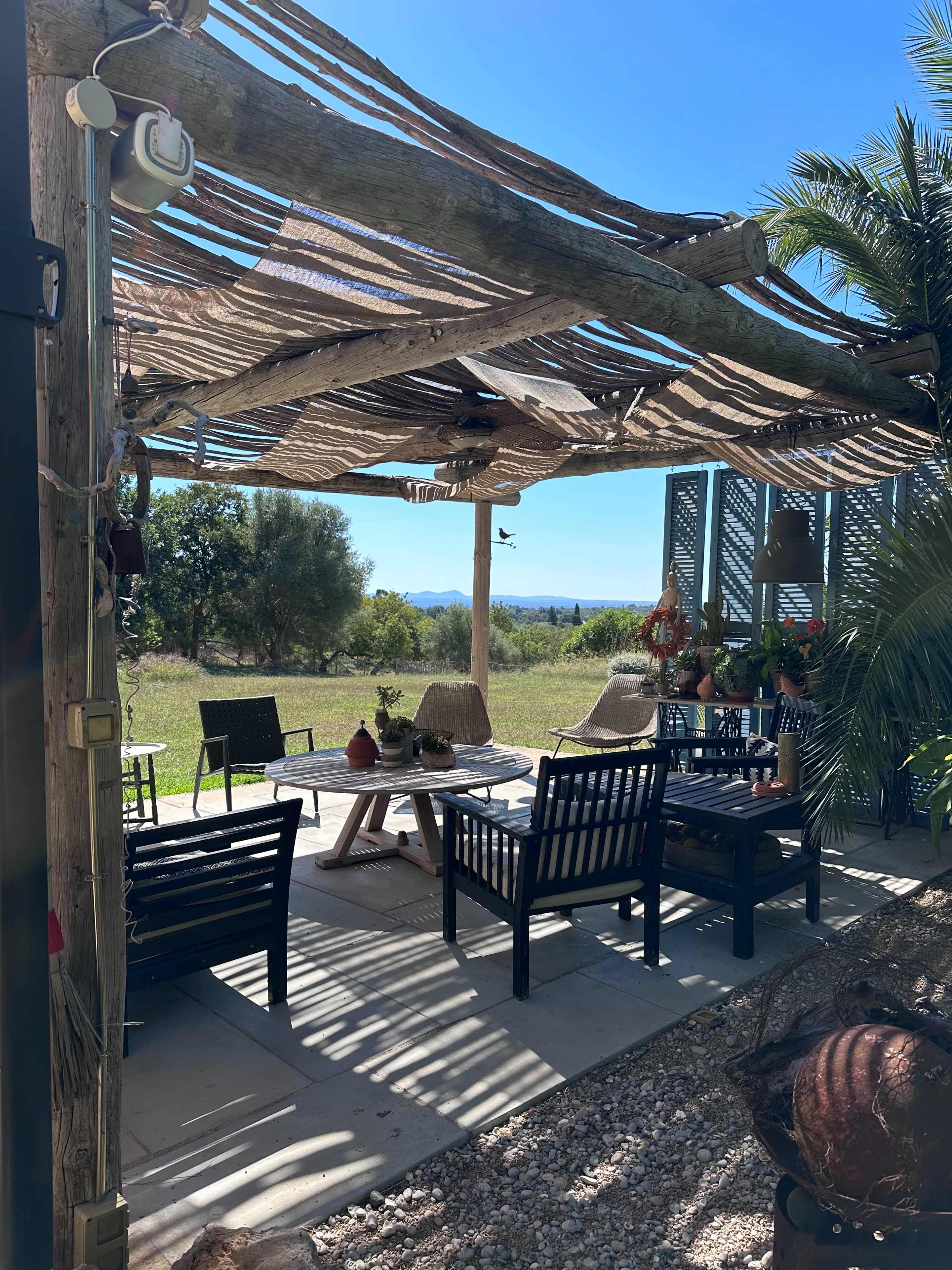 A shaded outdoor seating area features wooden chairs and a table, overlooking a grassy field and distant mountains.