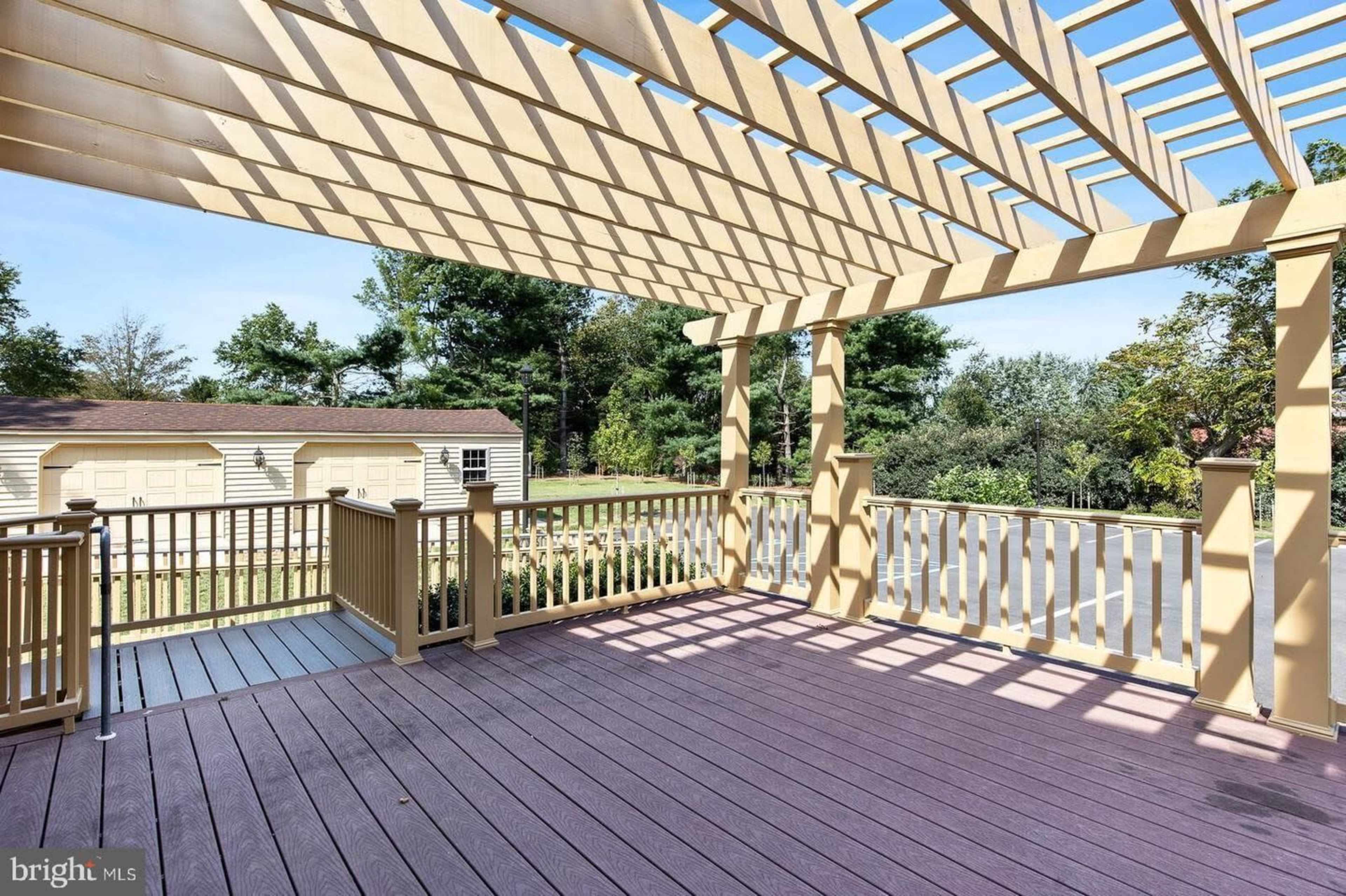 A wooden deck with a pergola structure above, surrounded by trees and a nearby shed in the background.