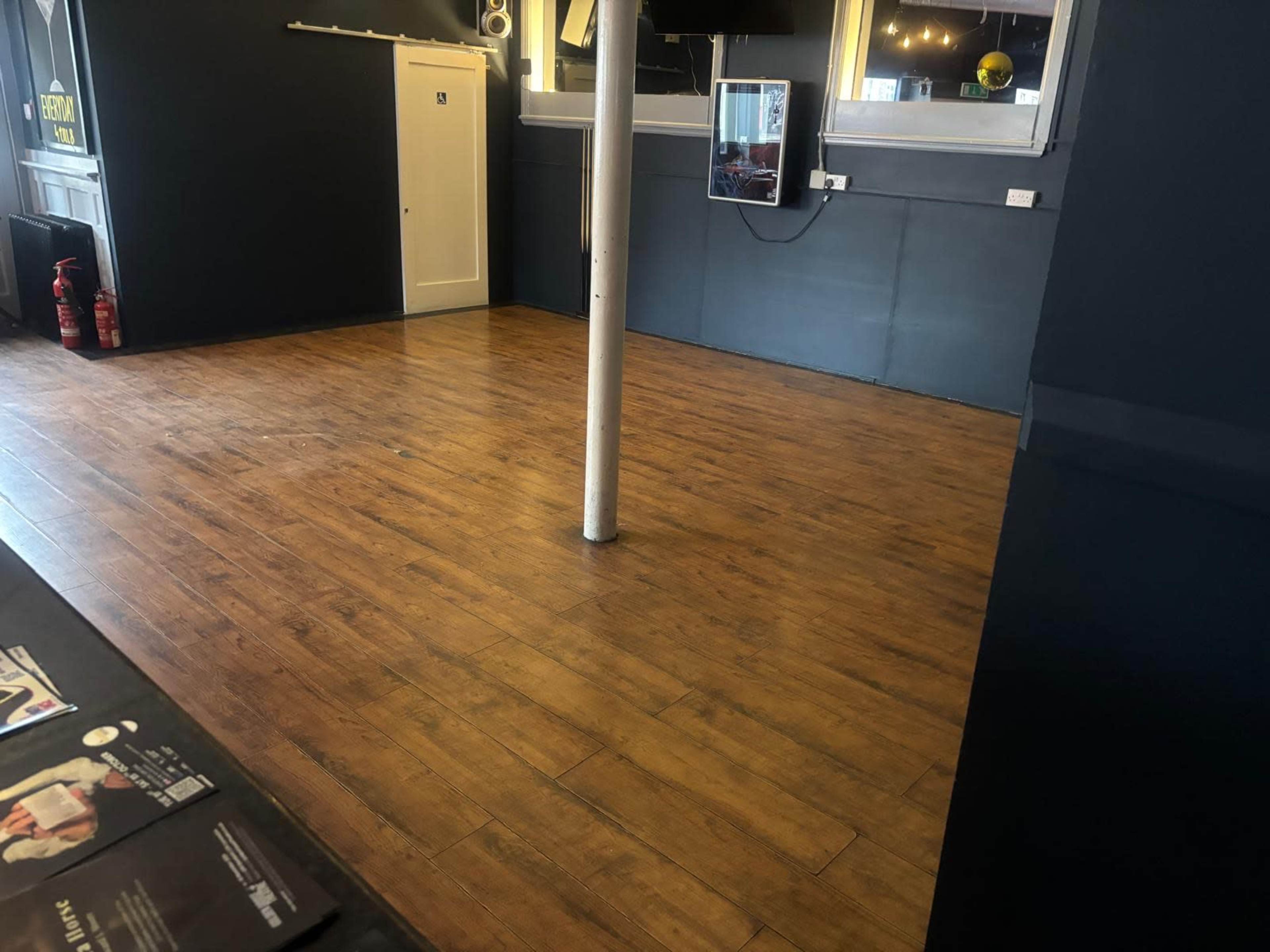 The image shows an empty room with wooden floors, a white door, a wall-mounted mirror, and a central support pole.