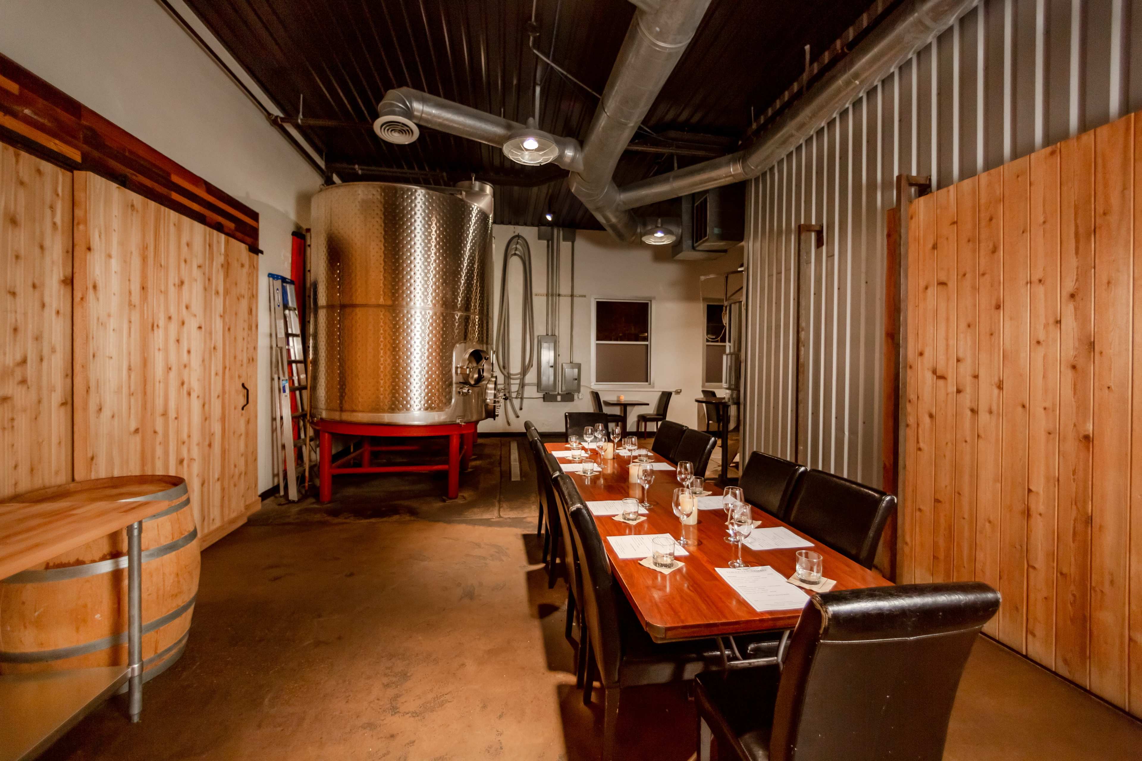 A dining area with a long wooden table set for a meal, adjacent to a large metal fermentation tank and wooden walls.