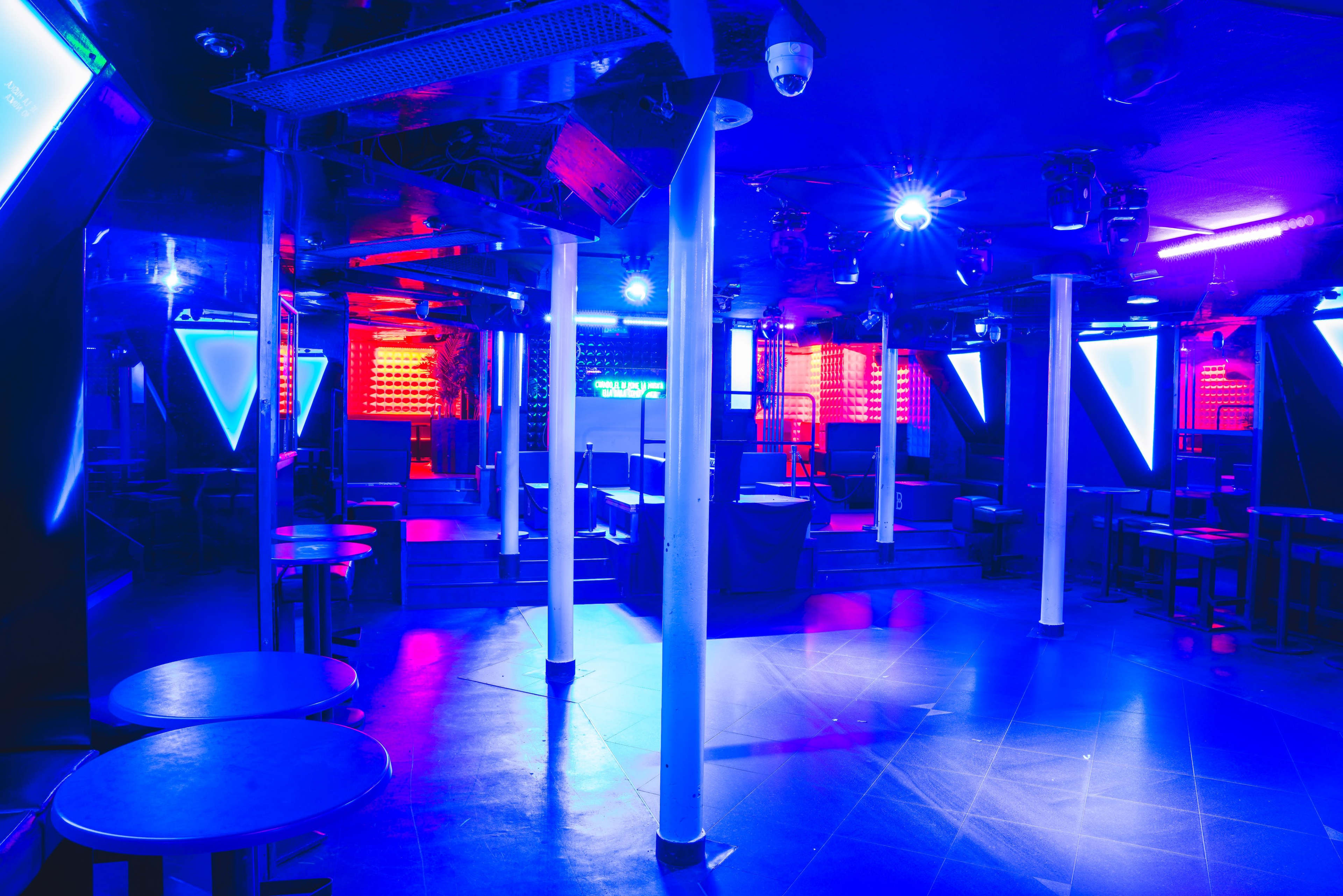 The image shows an empty nightclub interior with blue and red lighting, featuring tables and a dance floor.