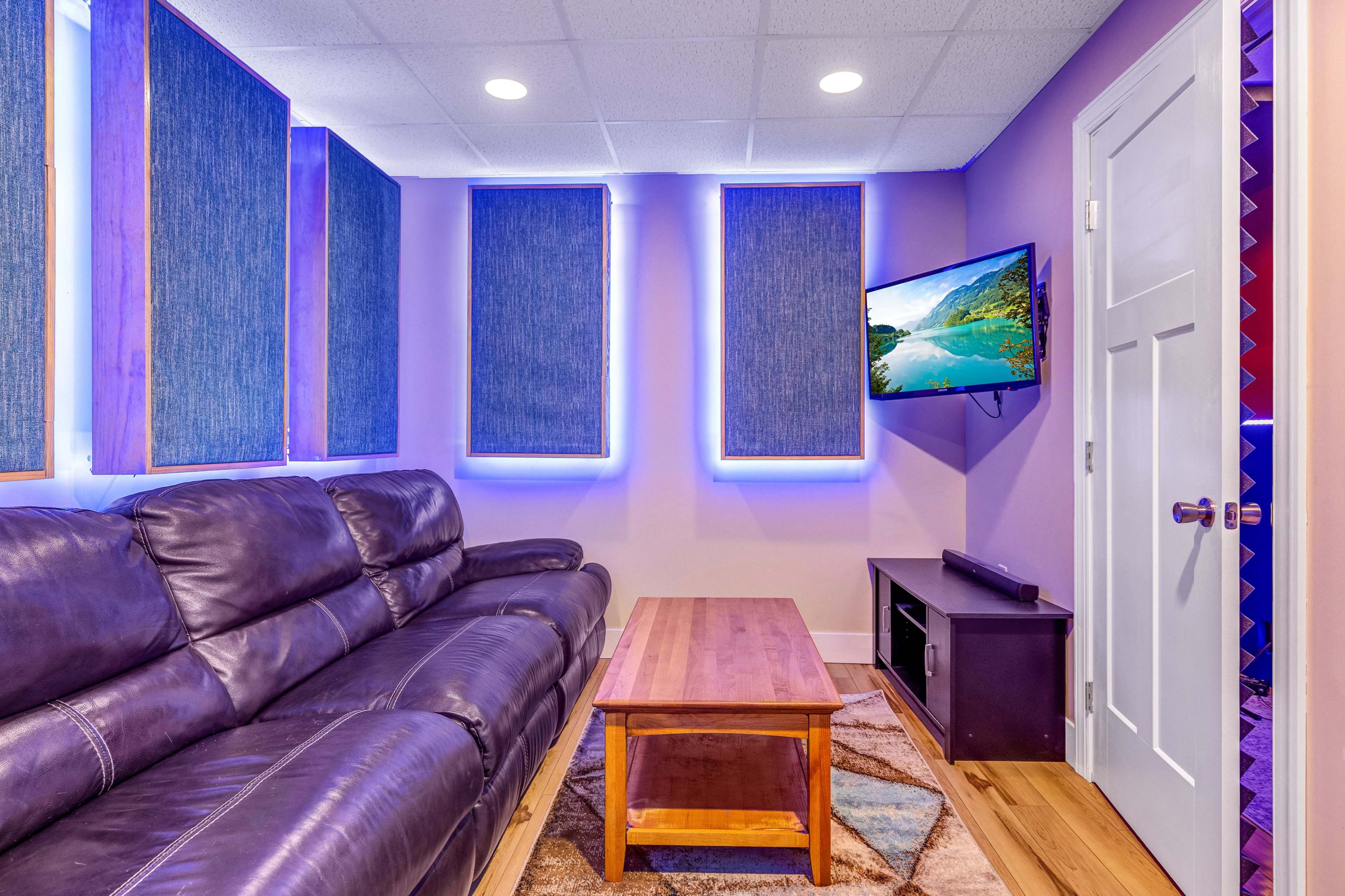 A cozy media room features a leather couch, a wooden coffee table, and a mounted television on a purple wall with illuminated window treatments.