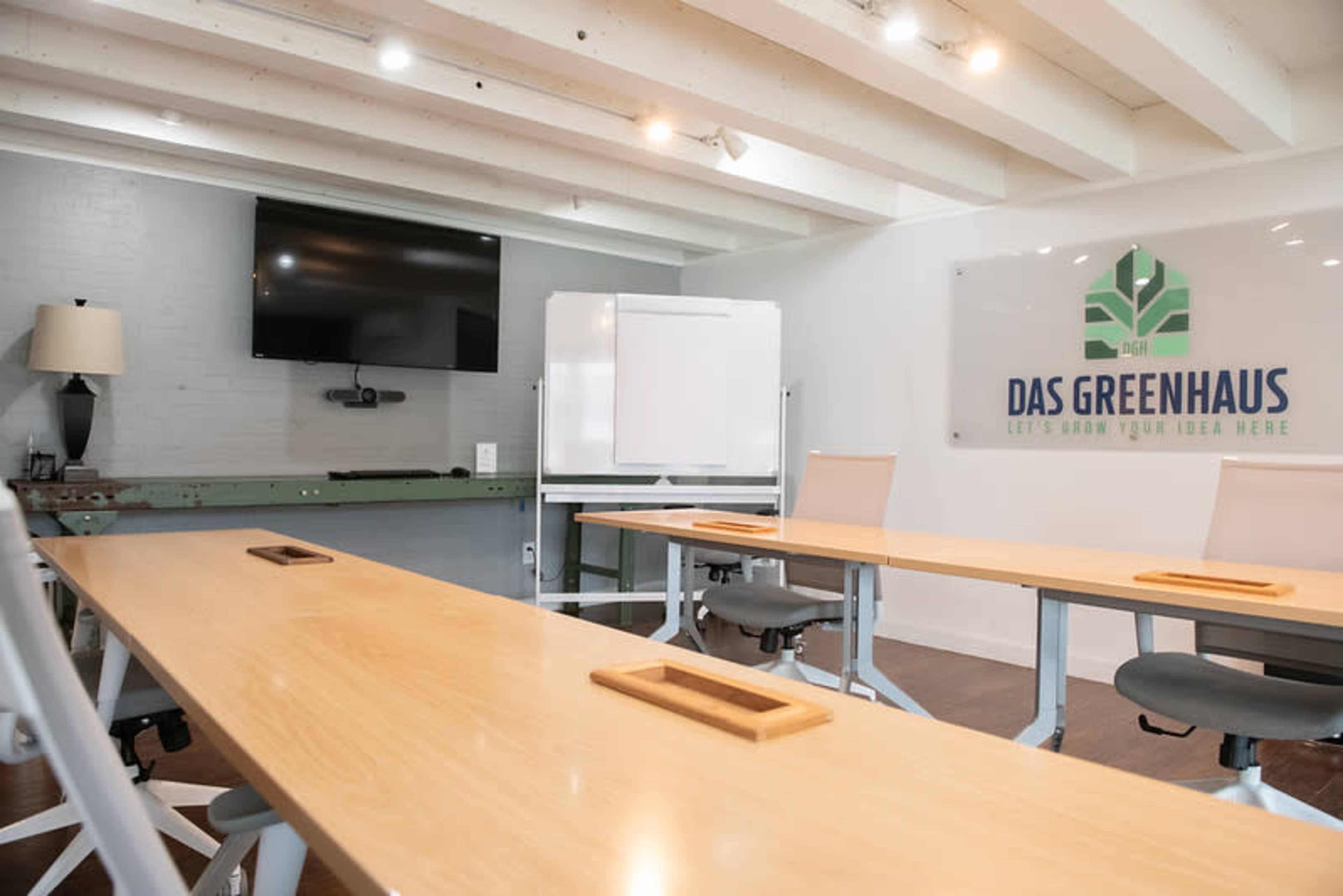 The image shows a modern conference room with light wood tables, office chairs, a wall-mounted TV, a whiteboard, and a sign that reads "DAS GREENHAUS."
