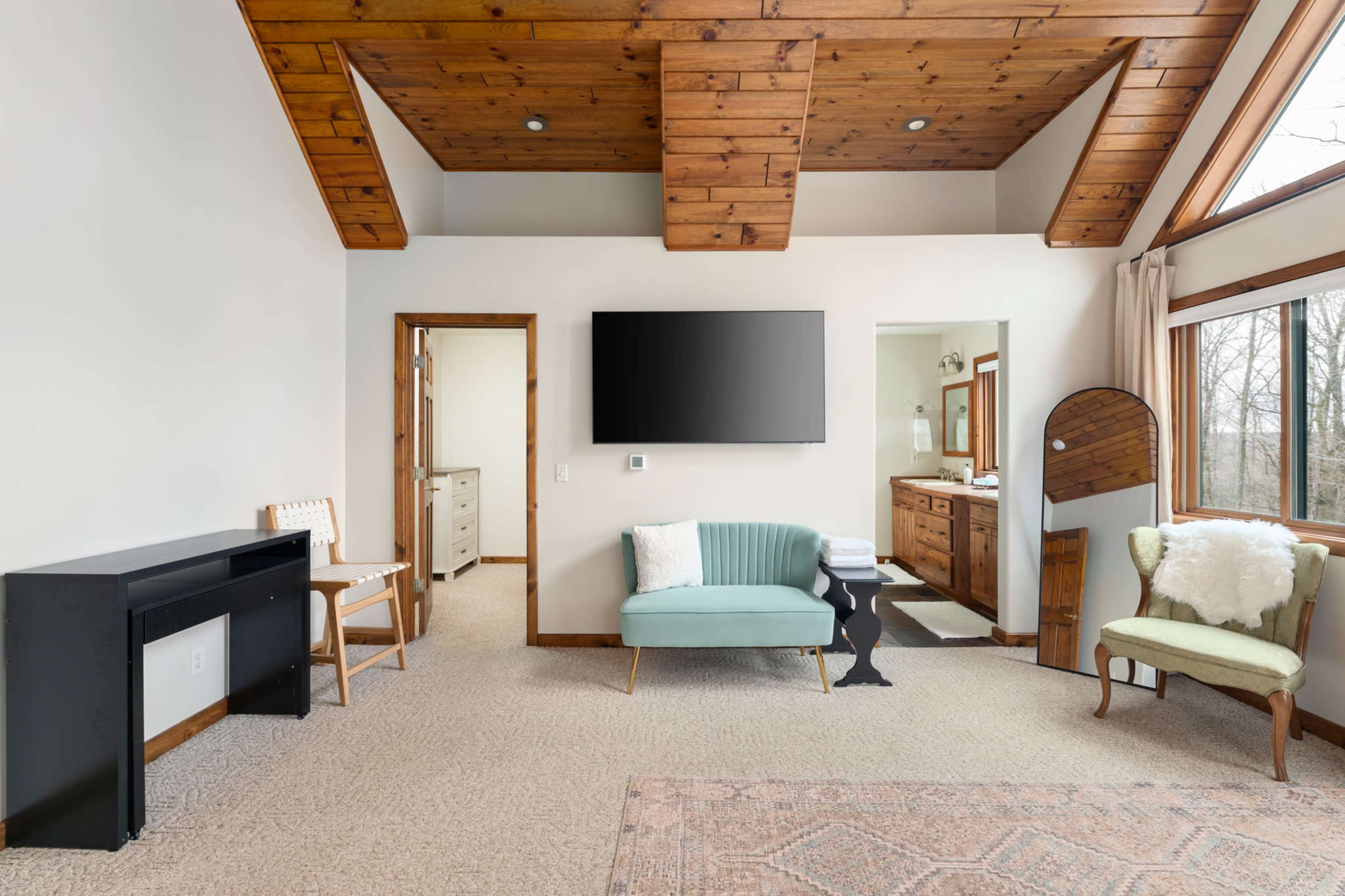 The image shows a spacious, modern room featuring a green couch, a black desk, a large television mounted on the wall, and an ensuite bathroom visible in the background.
