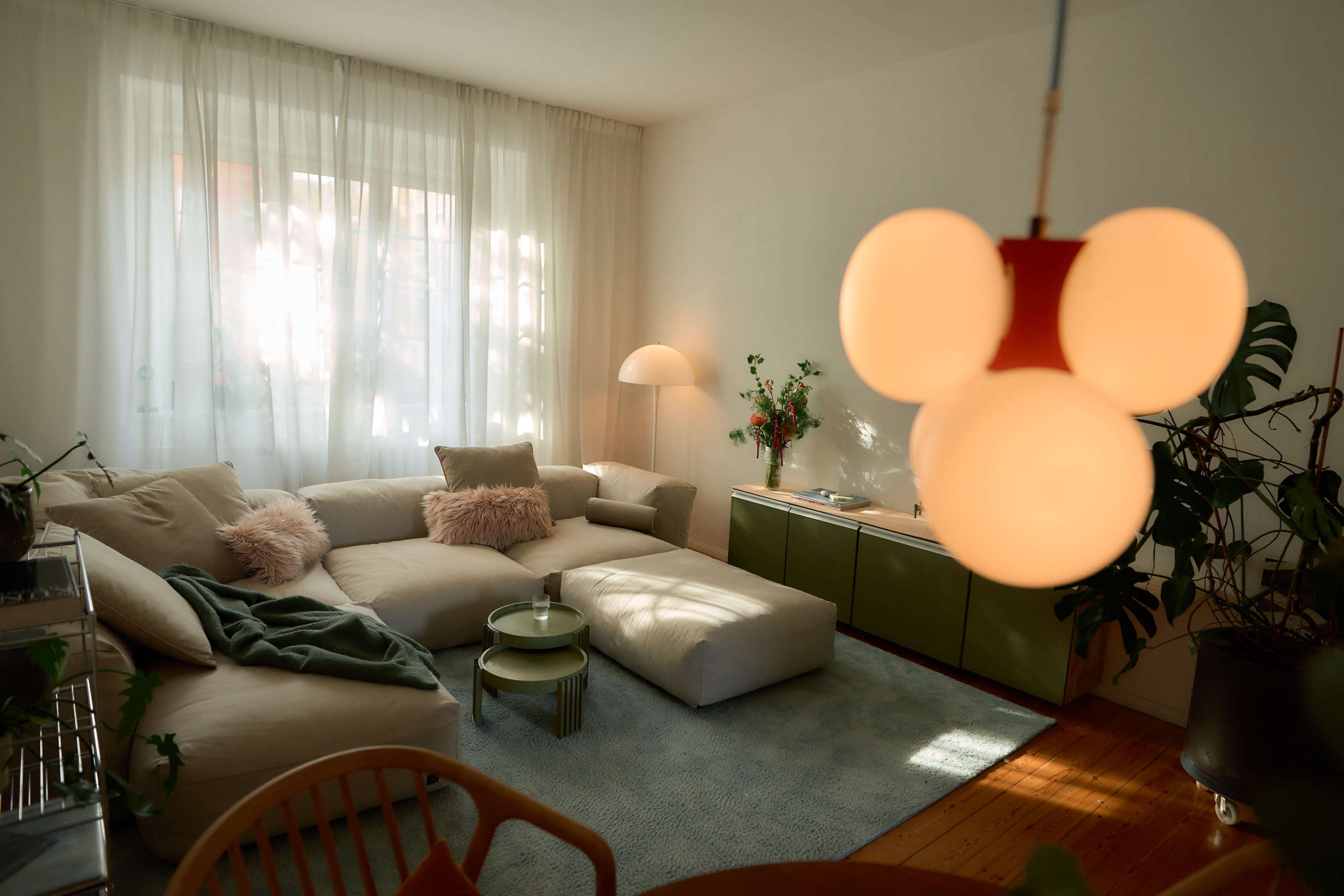 The living room features a large sectional sofa, a minimalist coffee table, and a soft rug, complemented by natural light streaming in through sheer curtains.