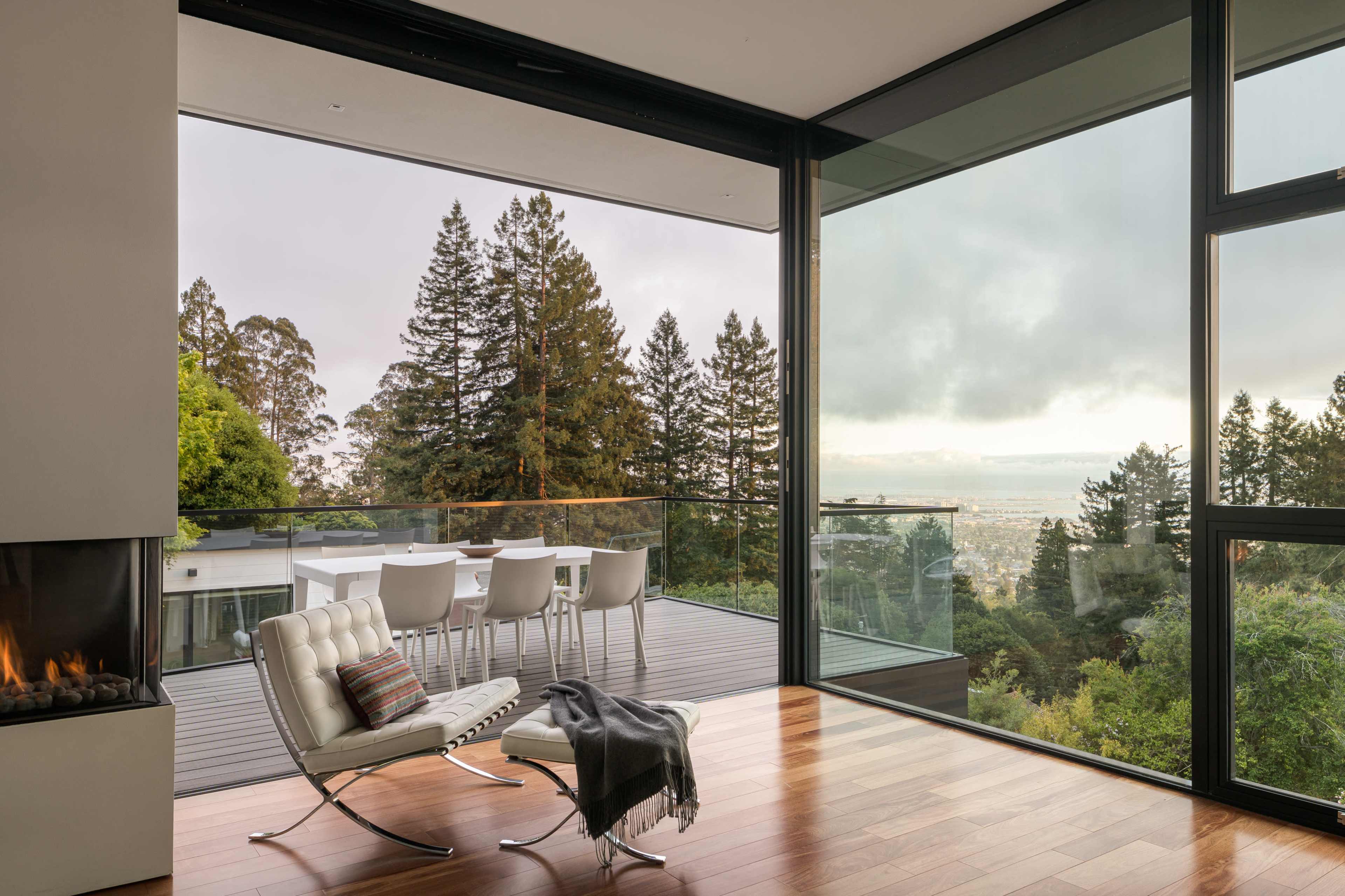 A modern living room features a glass wall that opens to a terrace with outdoor seating and a view of a forested landscape.