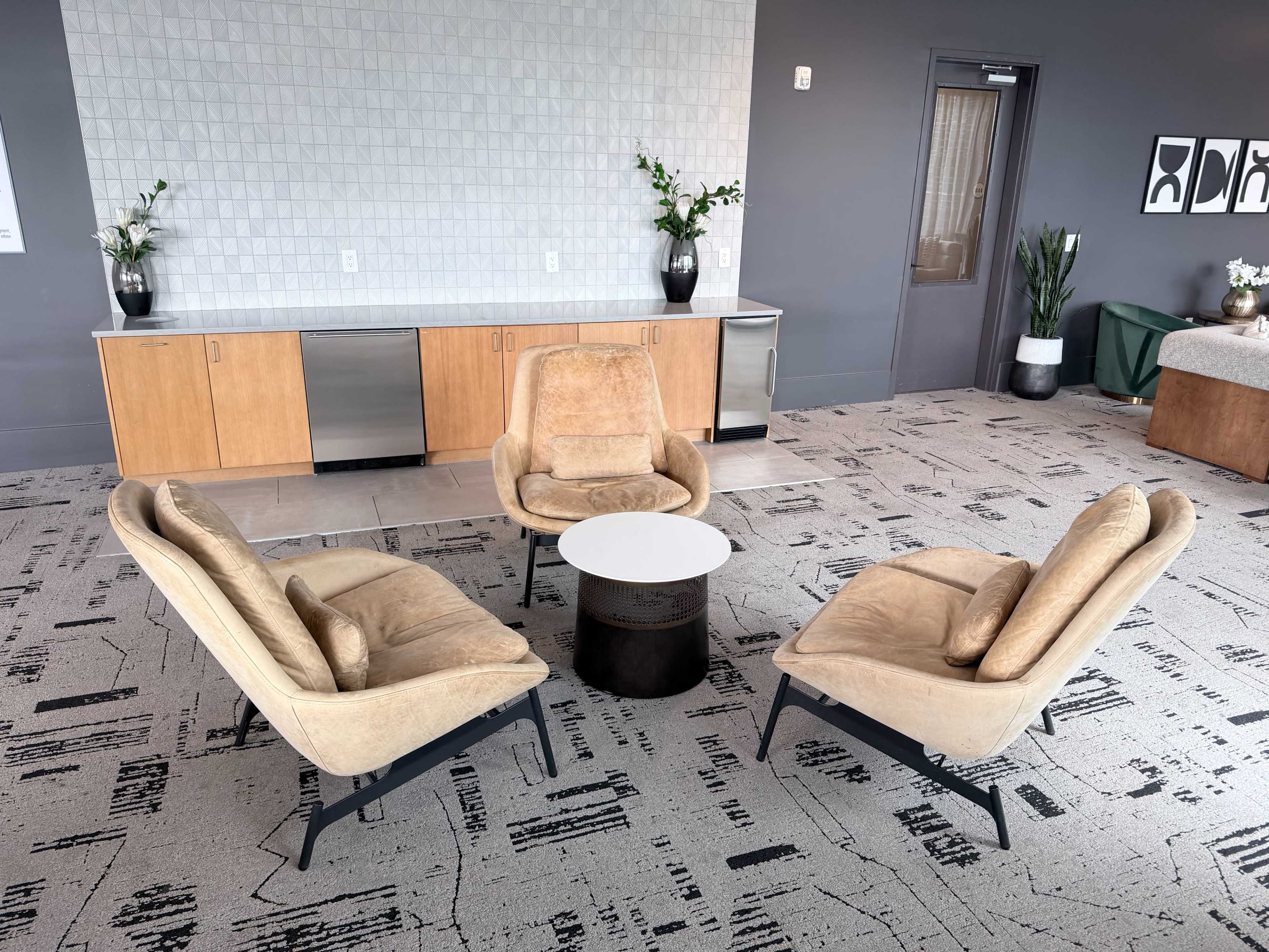 The image features a modern lounge area with three beige armchairs arranged around a circular white table on a textured carpet, set against a backdrop of a sleek gray wall and cabinetry.