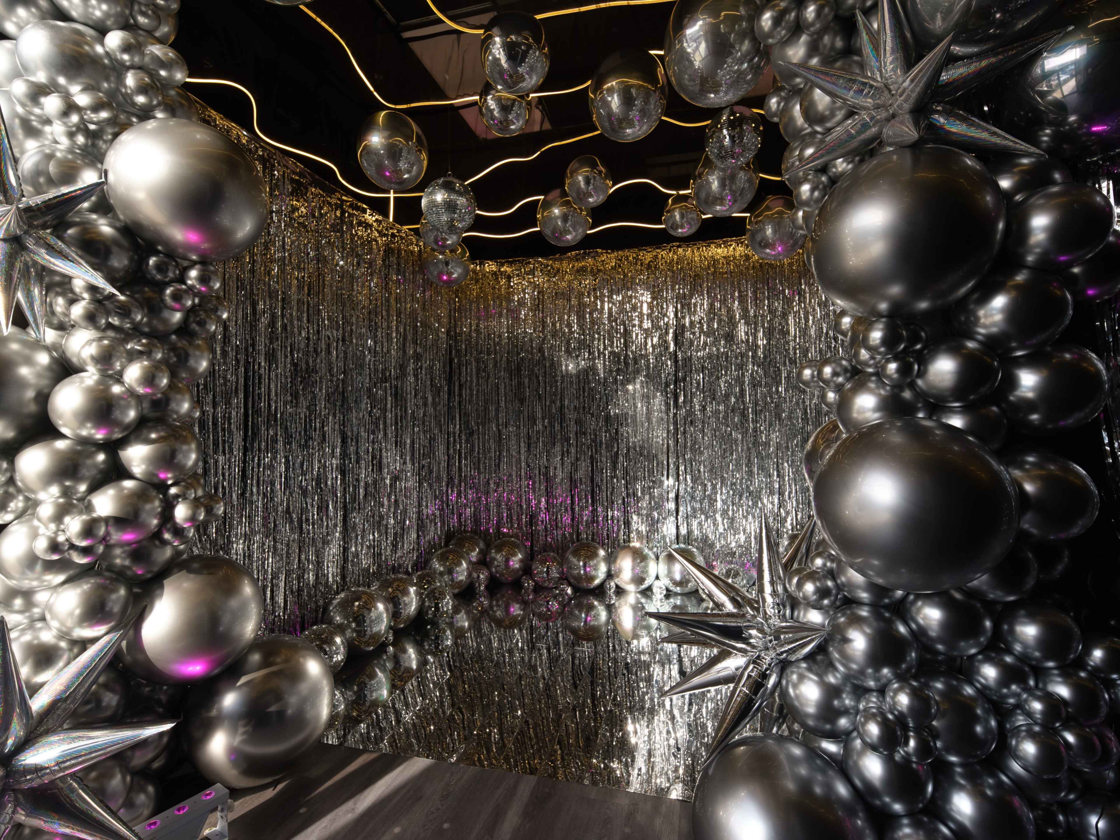 The image shows a room decorated with an array of metallic balloons and shiny silver fringe, creating a festive atmosphere.