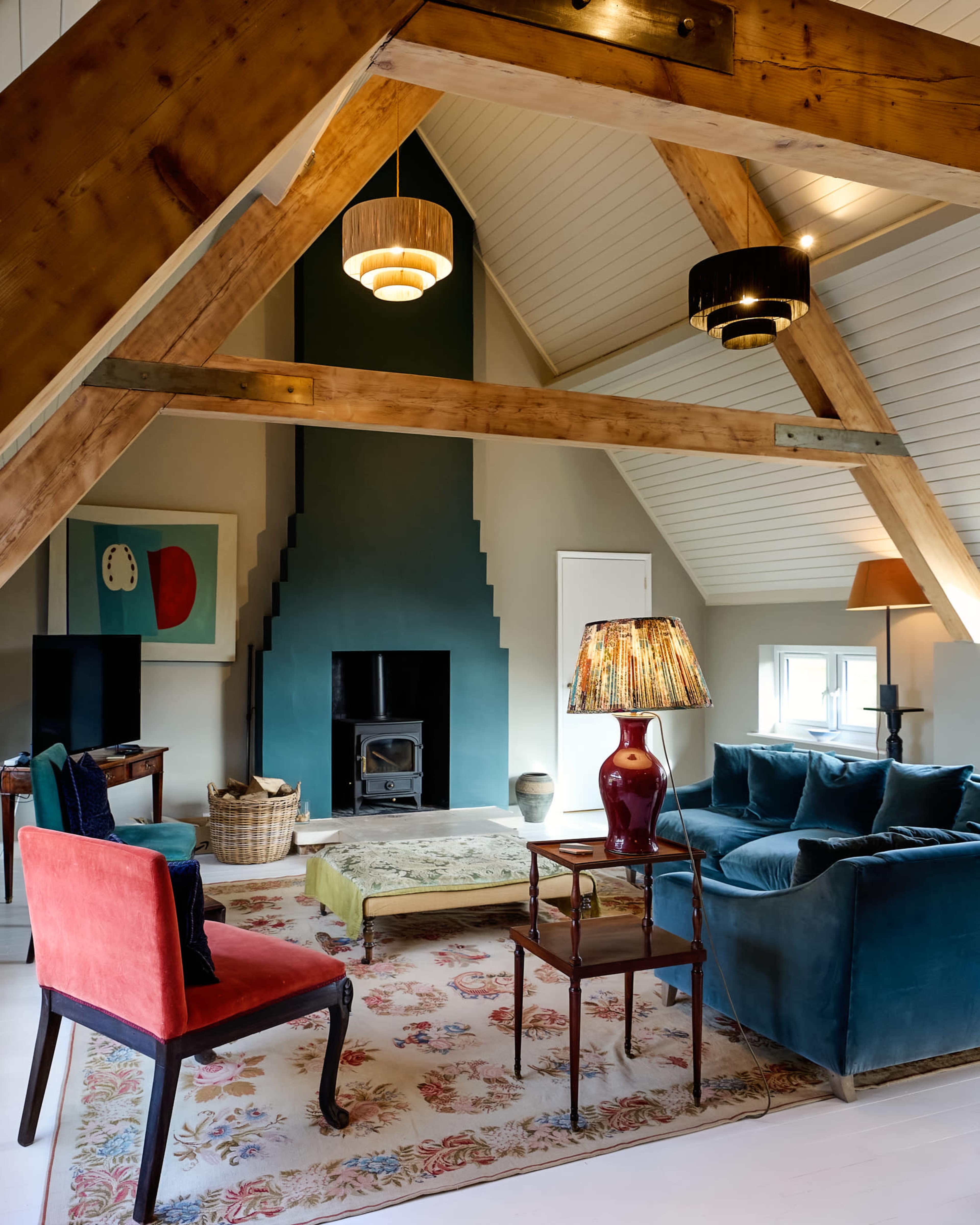 The image shows a cozy attic living space featuring a blue velvet sofa, a patterned area rug, wooden beams, and a modern fireplace.