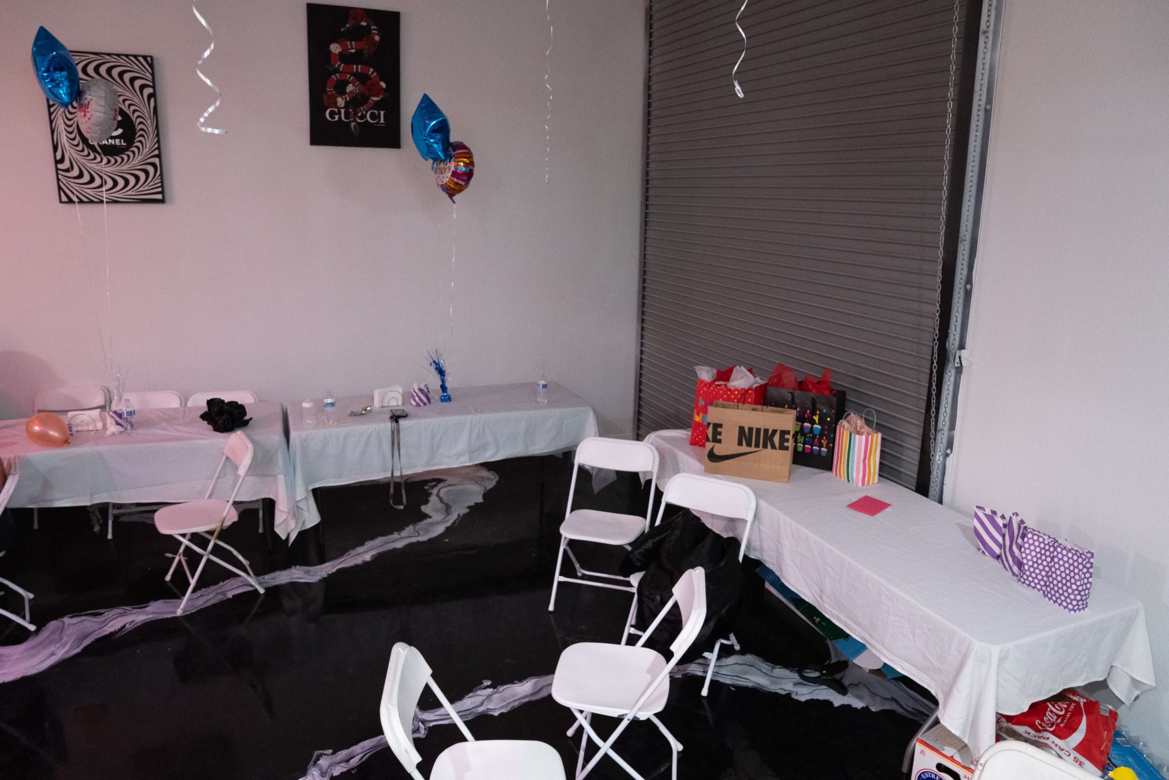 A party setup with white tables and chairs, decorated with balloons, and gifts stored on a shelf against a wall.