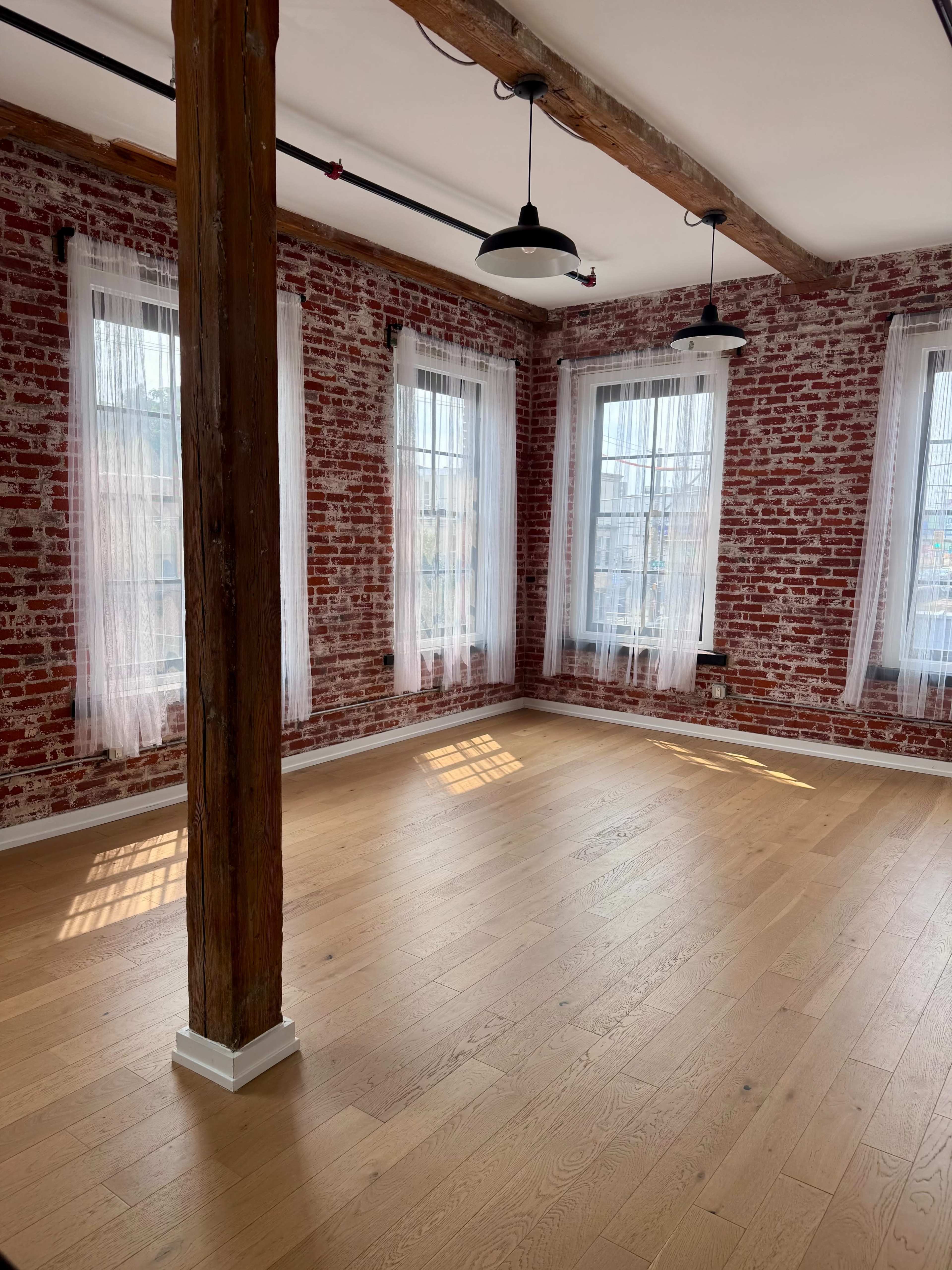 The image shows a bright room with exposed brick walls, large windows with sheer curtains, and wooden beams, featuring a wooden floor.