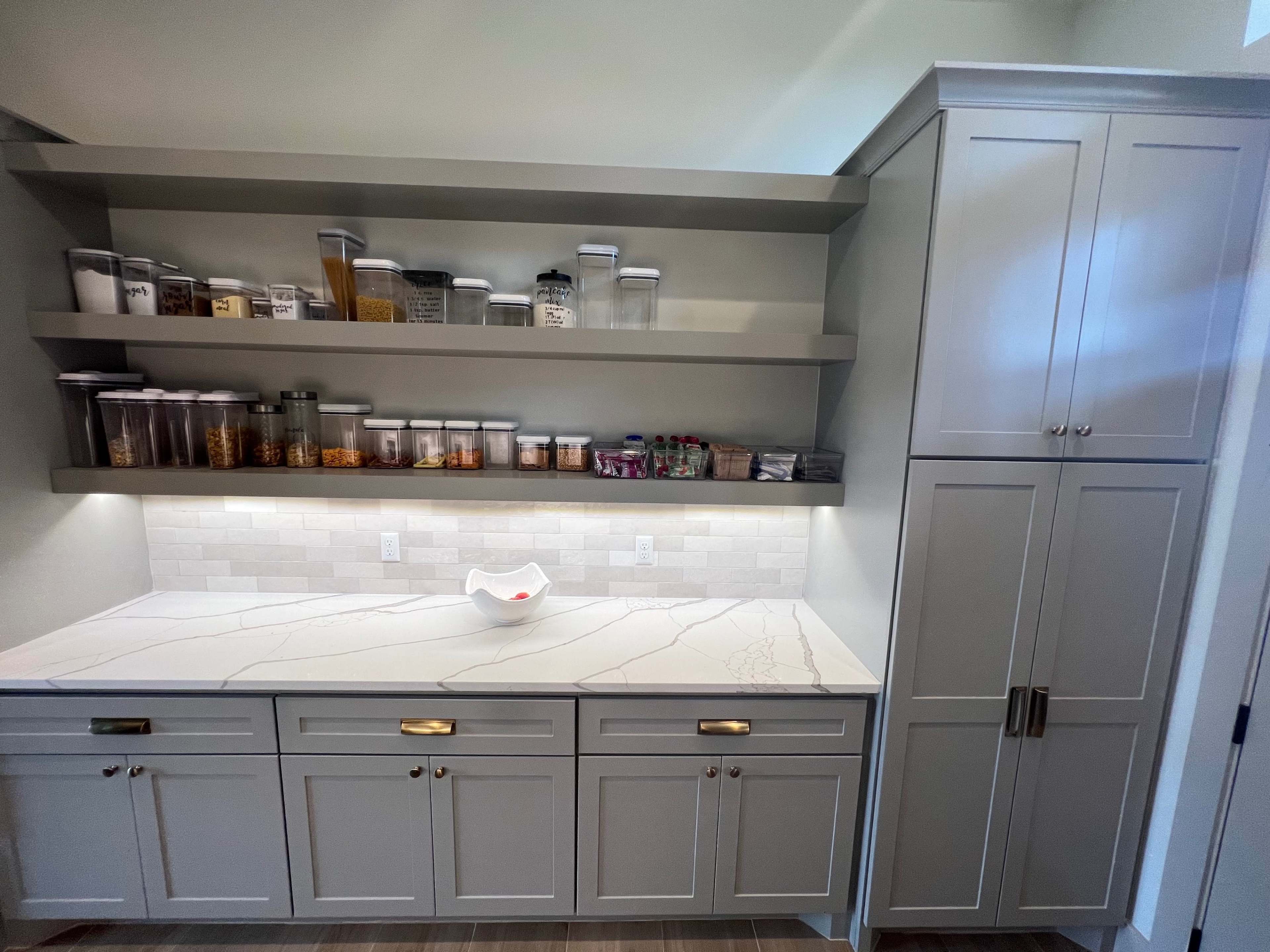 A modern kitchen pantry with two shelves of clear containers filled with various dry goods and a countertop beneath, featuring a light-colored stone surface and storage cabinets.