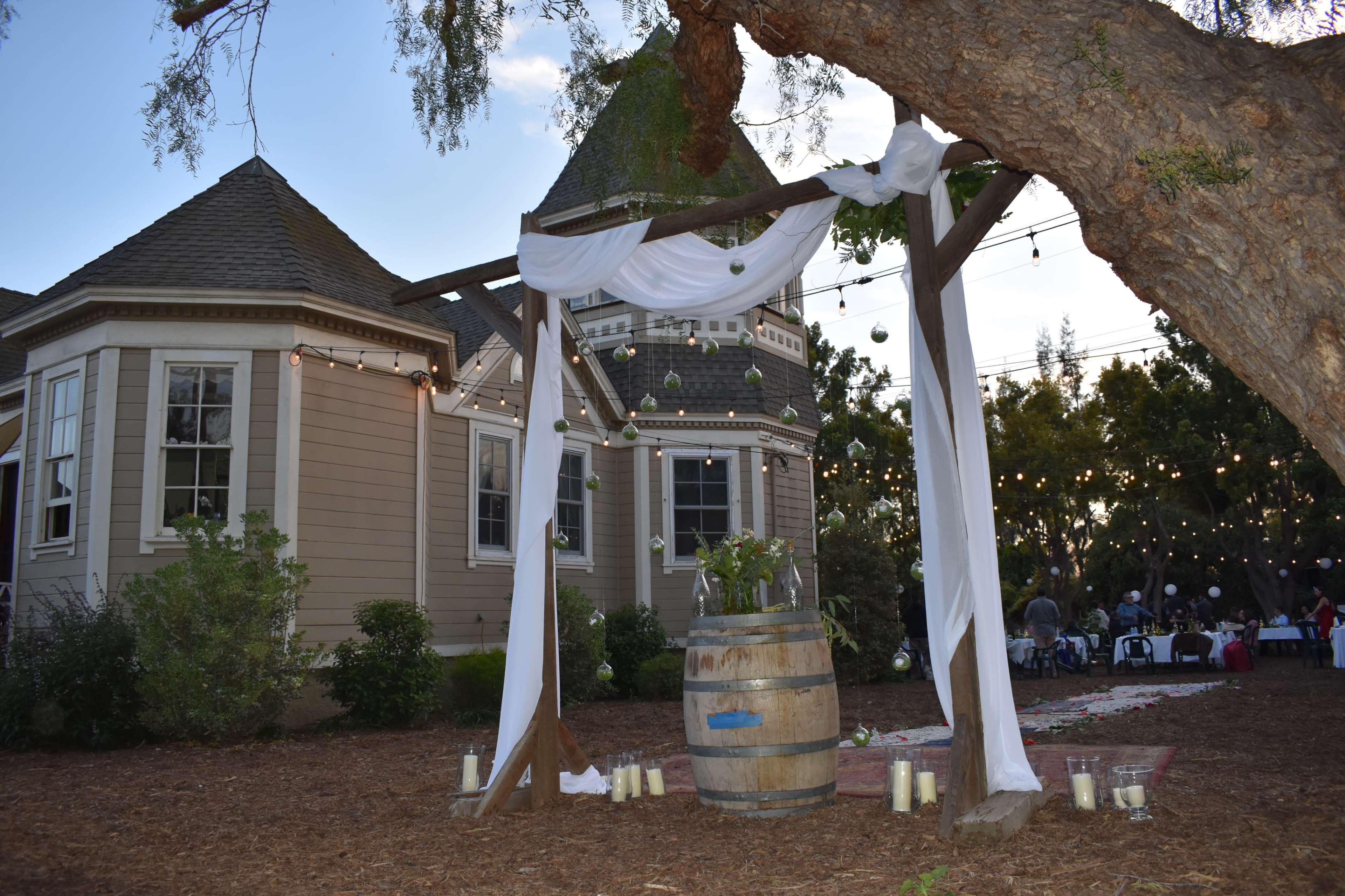 An outdoor wedding setup features a wooden arch draped with fabric, standing beside a large barrel, with string lights illuminating the scene behind a quaint house.