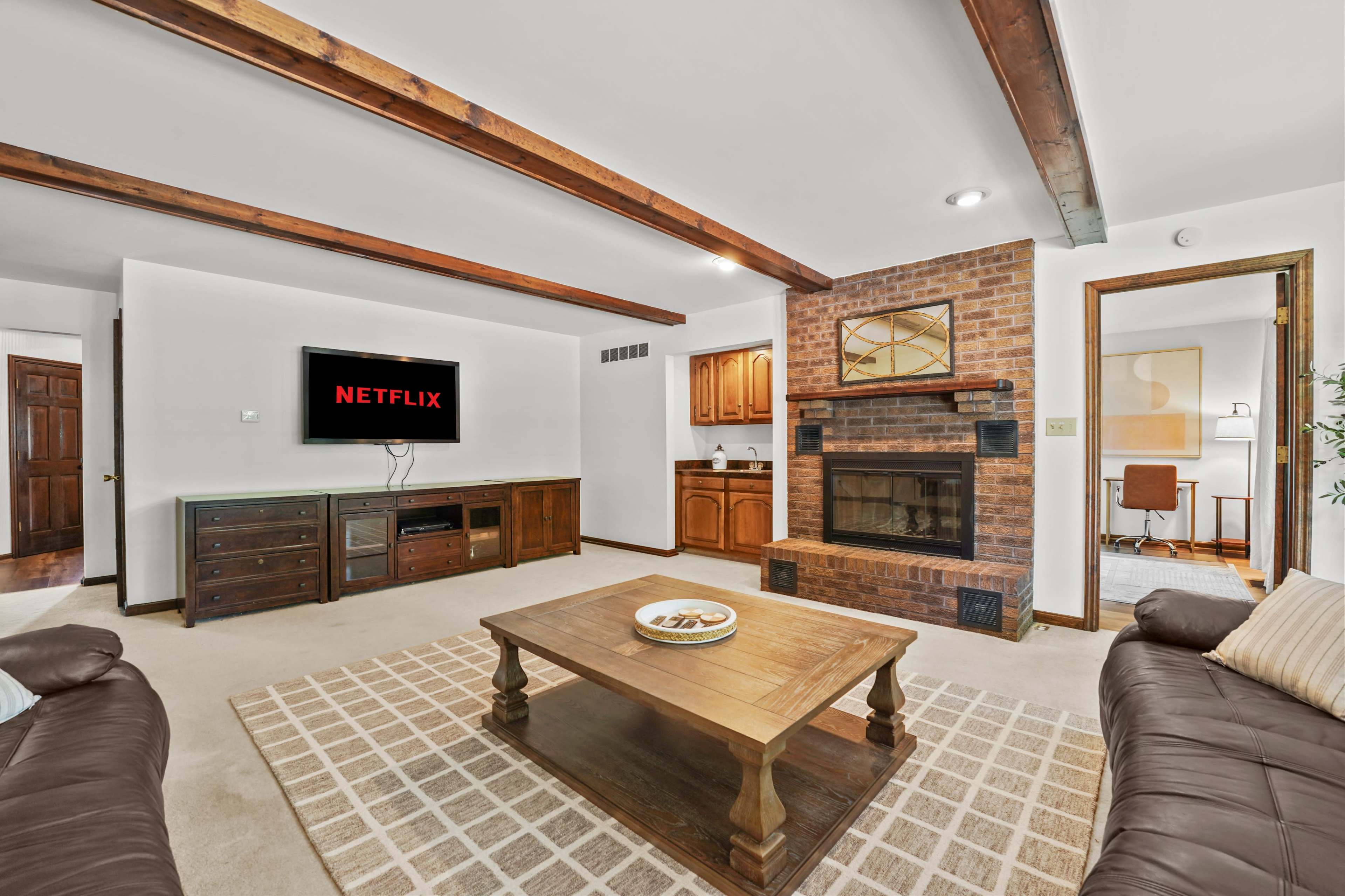 The living room features a brick fireplace, a coffee table, and a large television displaying the Netflix logo, with wooden beams on the ceiling.