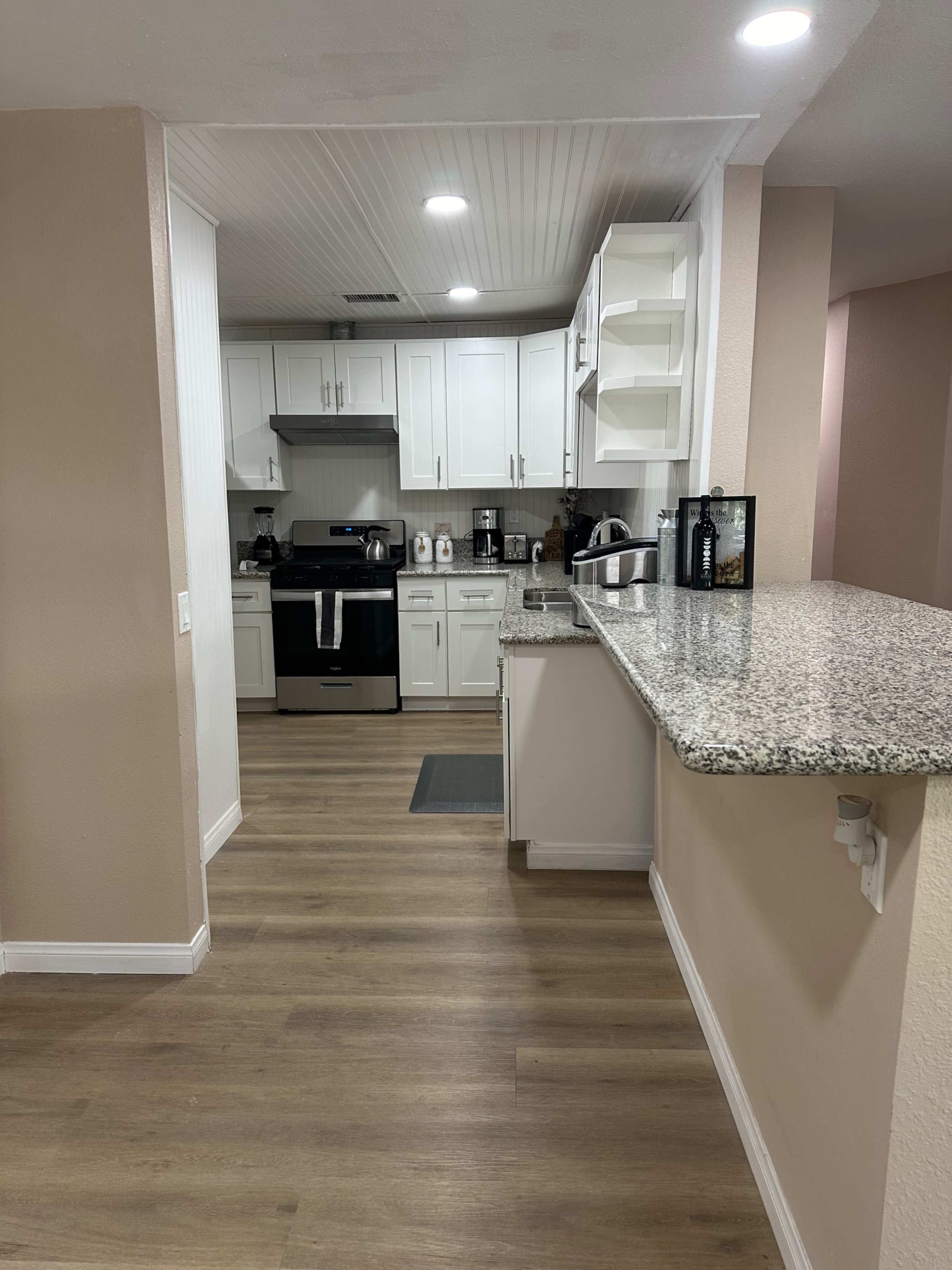 The image shows a modern kitchen with white cabinets, granite countertops, and wooden flooring, featuring an open layout that connects to a living space.