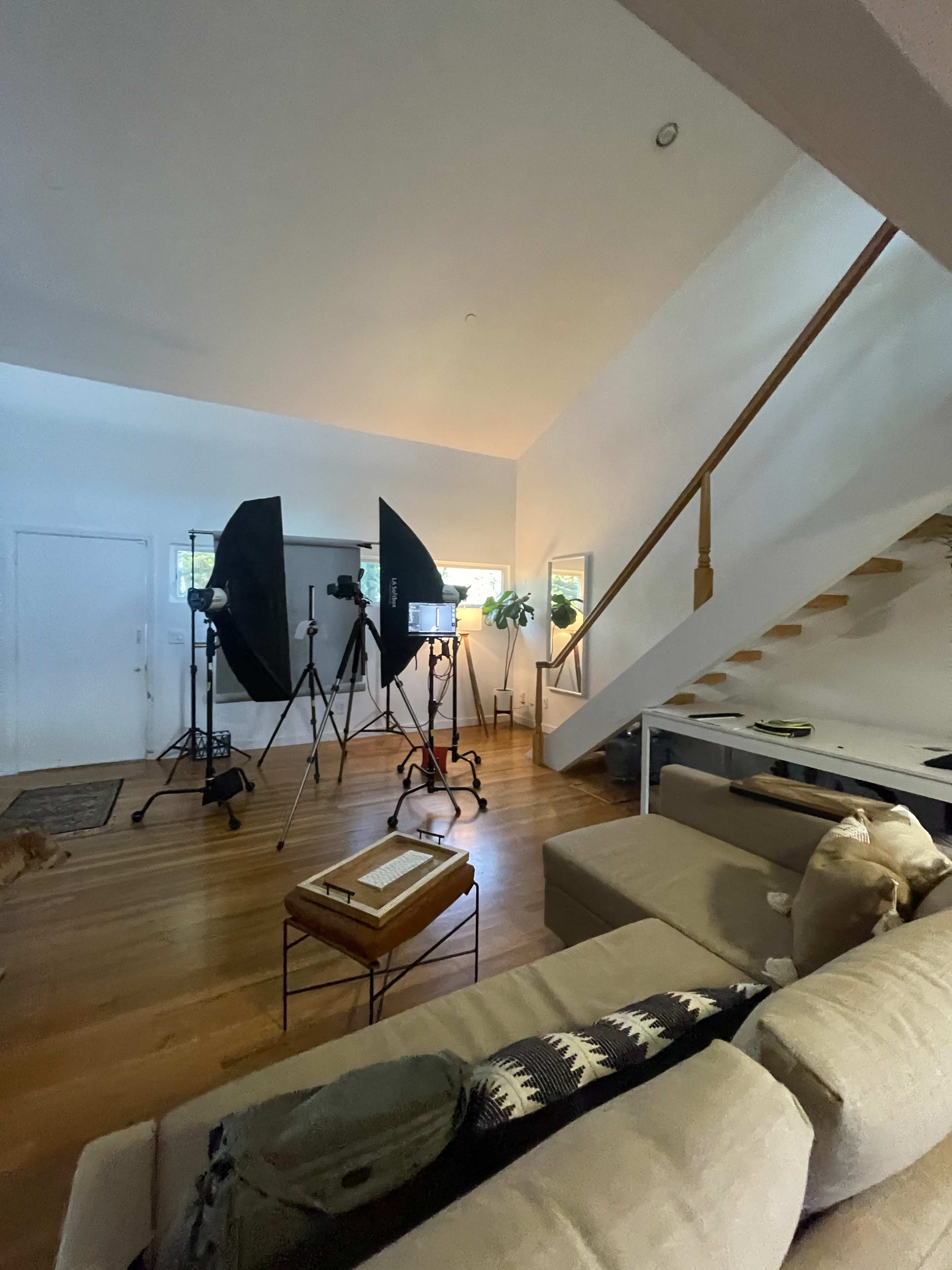 The image shows a brightly lit living room with video equipment set up, including tripods and softboxes, alongside a staircase and a sofa.