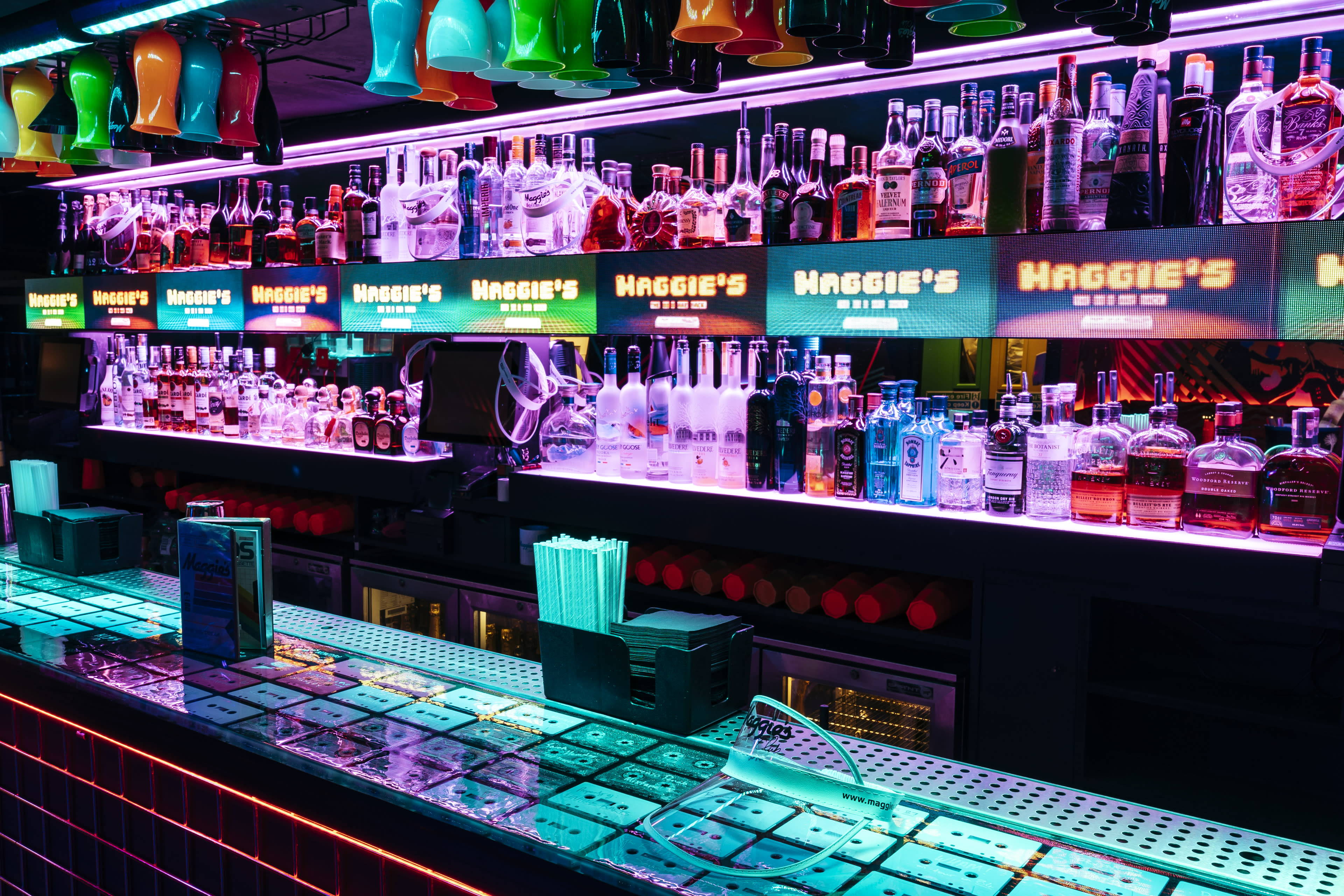 A brightly lit bar displays an array of colorful bottles and neon signage.