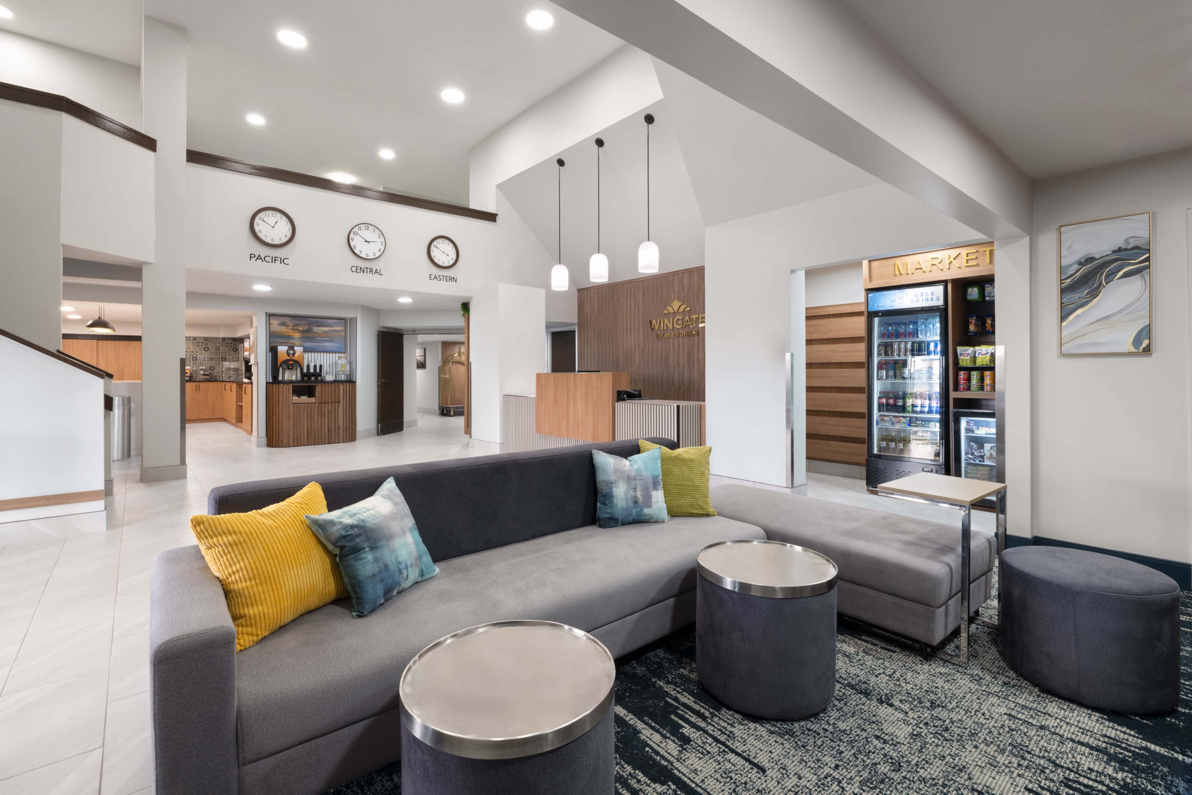 The image shows a modern hotel lobby featuring a curved gray sofa, round tables, and a reception desk, along with a small market area and clock displays on the wall.
