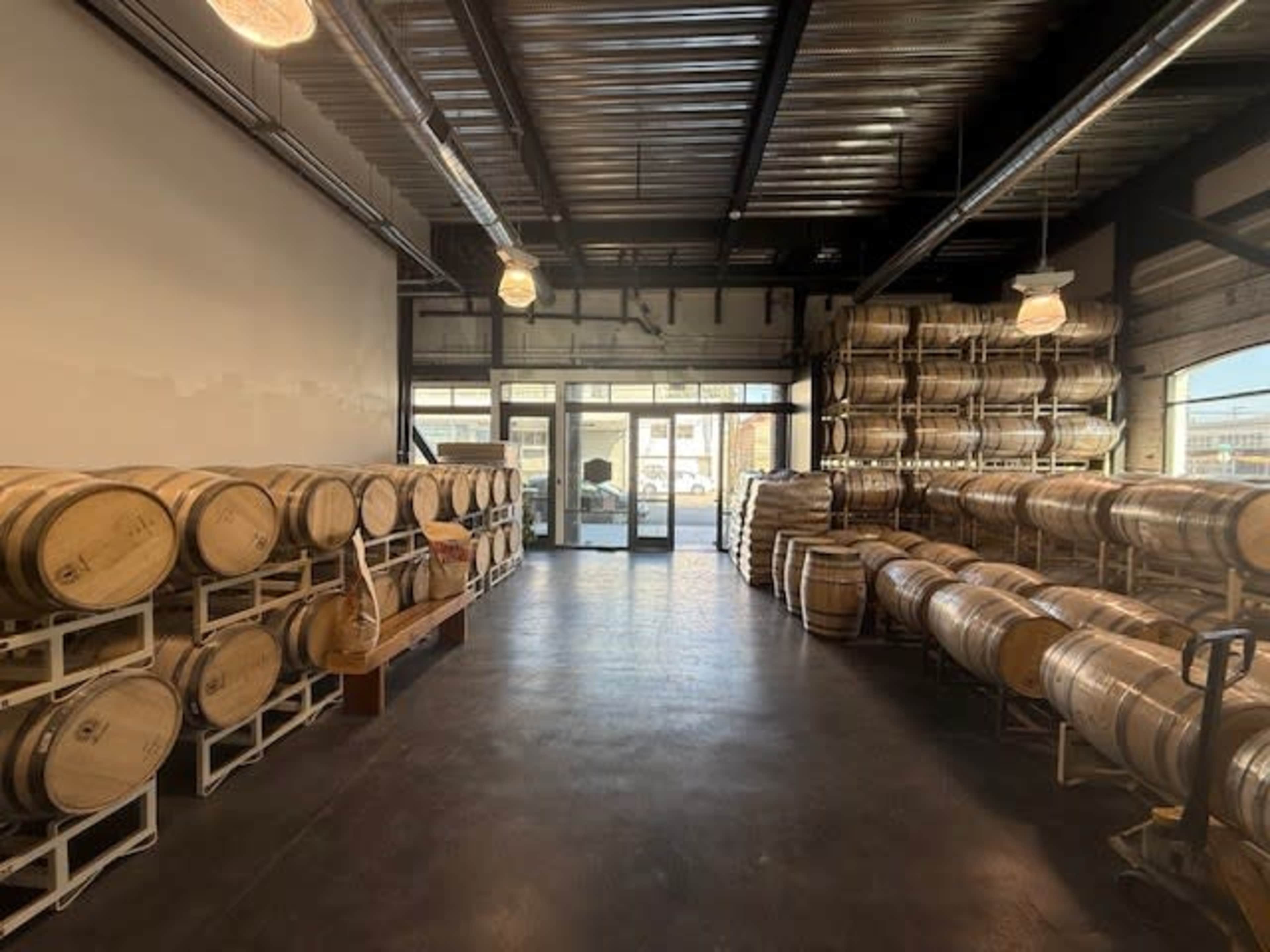 The interior of a winery showcases rows of wooden barrels stacked along the walls, with a large glass door leading outside.