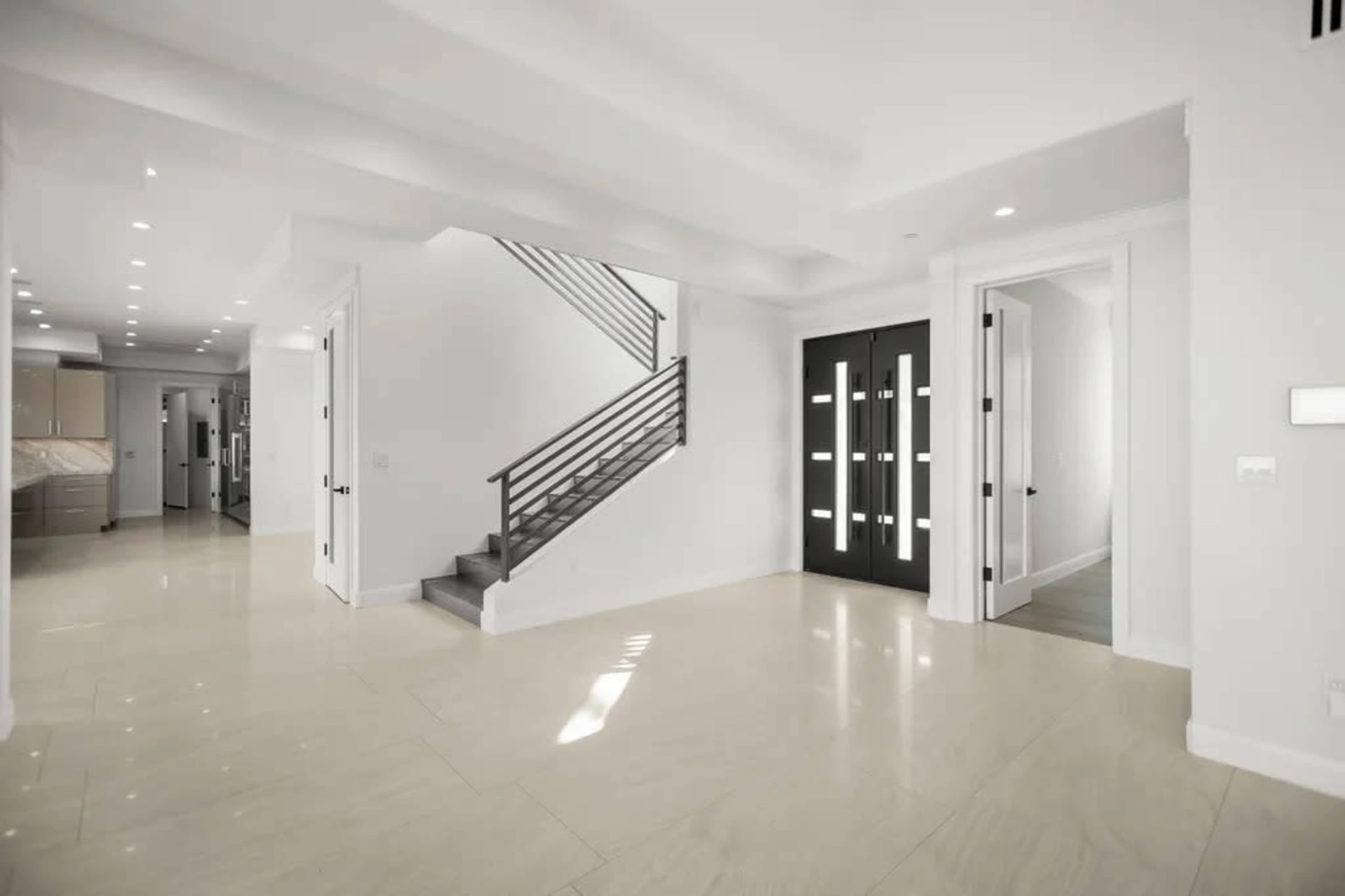 The image depicts a modern, spacious interior hallway with a staircase and a large front door featuring multiple vertical windows.