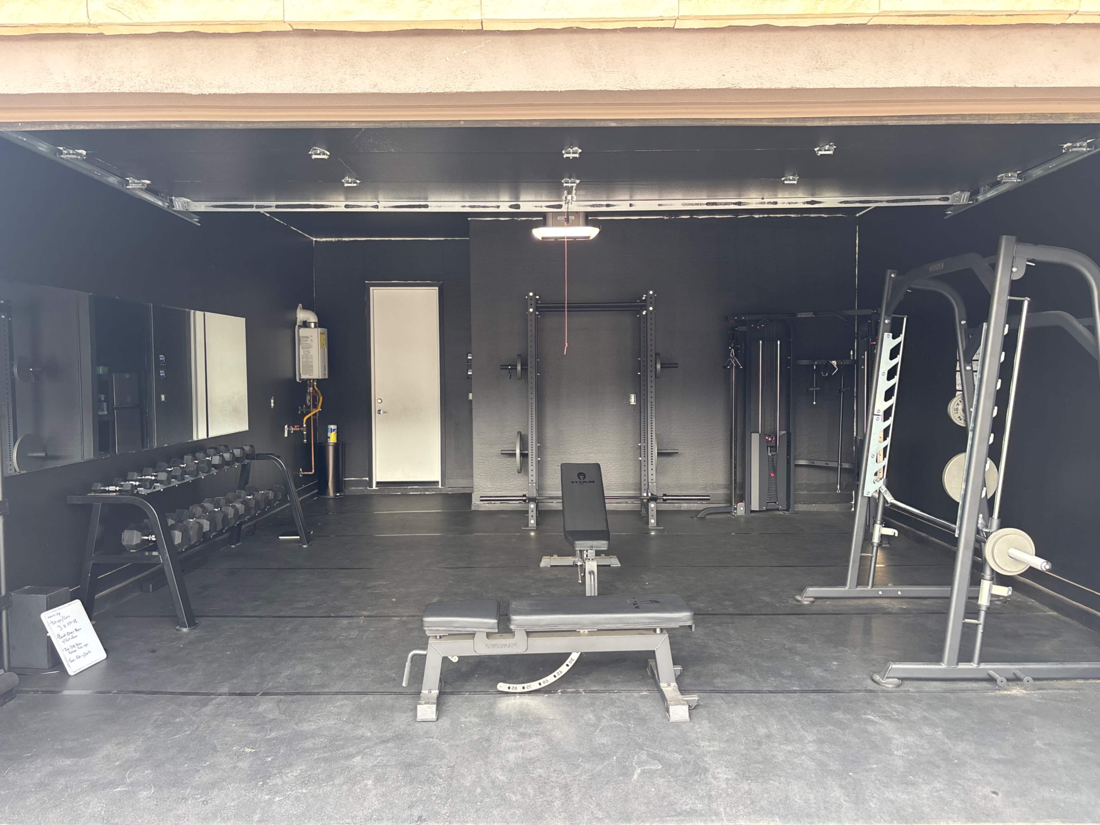 The image shows a home gym with a variety of weightlifting equipment, including dumbbells, a bench, and multi-use machines, all set within a garage space.