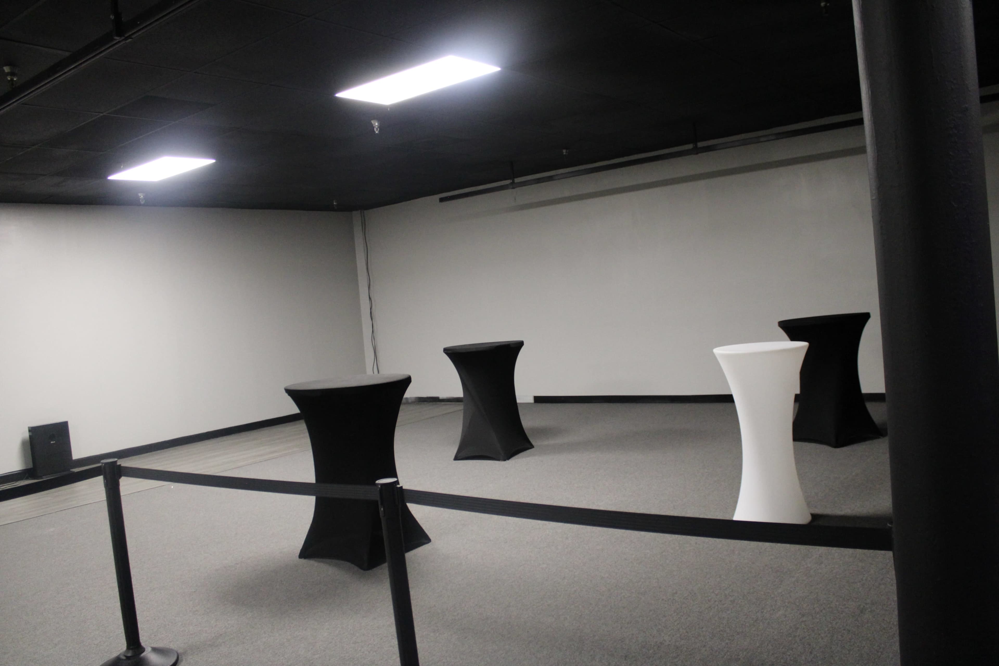 The image shows an empty room with gray carpet, featuring four tall tables covered in black and white cloth, surrounded by a black retractable barrier.